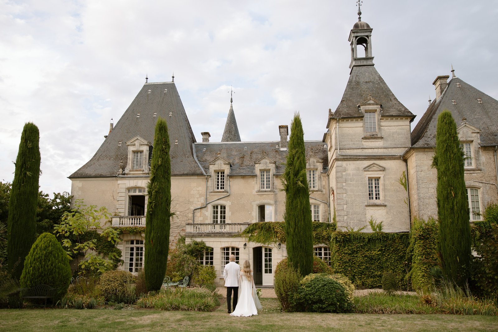 A couple in formal wedding attire stands hand in hand facing a large, historic chateau with tall pointed roofs and lush greenery.