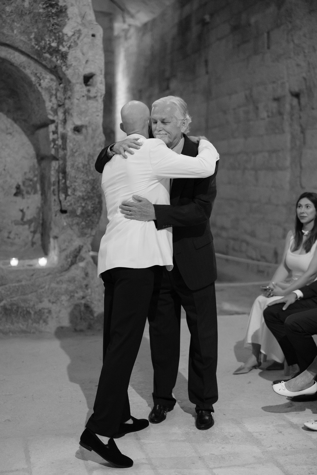 Two men embrace in a hug inside a stone-walled room, while a seated woman looks on. The scene is in black and white. Aubeterre sur dronne wedding.
