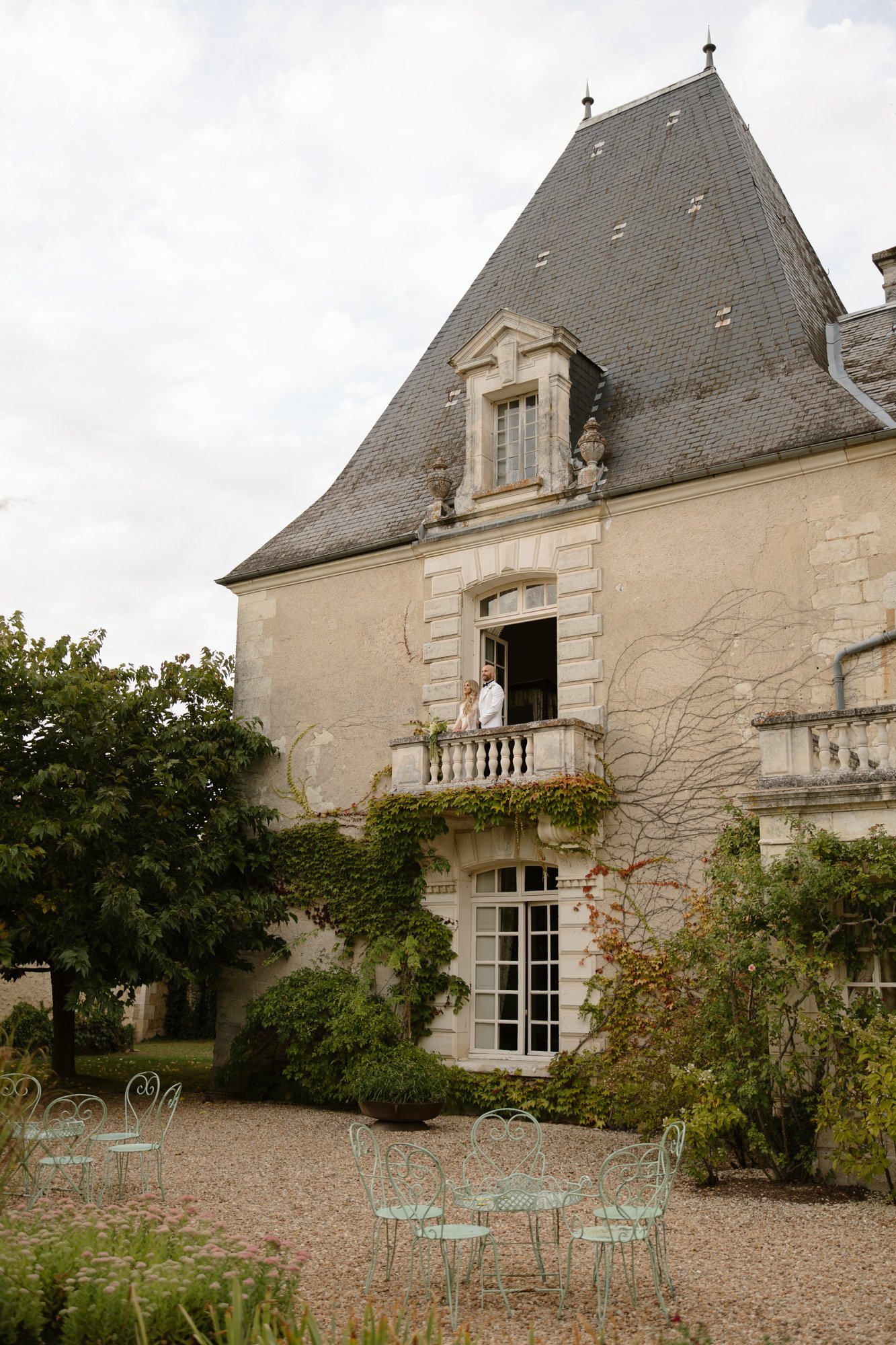 A person stands on a balcony of an old stone building with a steep slate roof, surrounded by climbing plants, trees, and metal garden furniture on a gravel patio.
