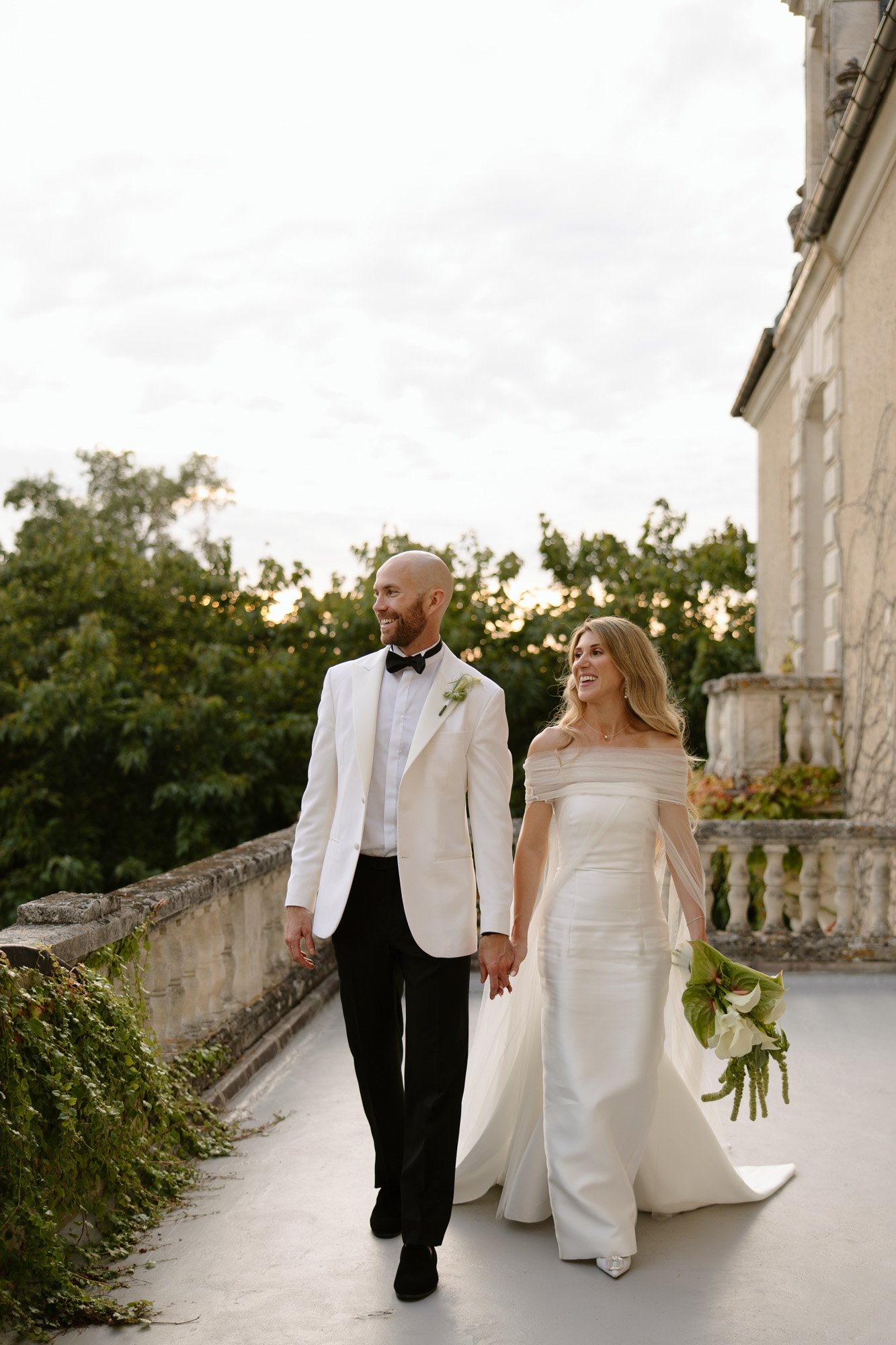 A bride and groom walk hand in hand outdoors, both dressed in formal wedding attire. The bride holds a bouquet and wears an off-shoulder gown; the groom wears a white jacket and black bow tie.