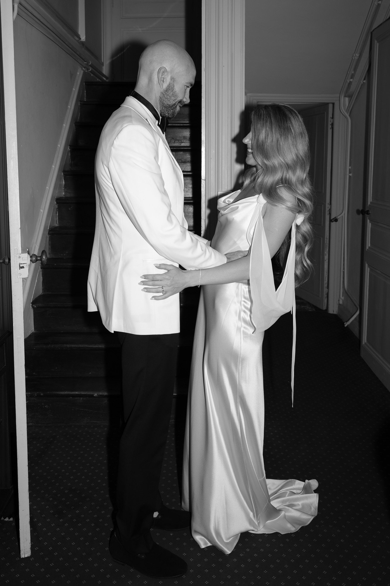 A man and woman in formal attire stand facing each other, holding hands, in front of a staircase in an indoor setting.