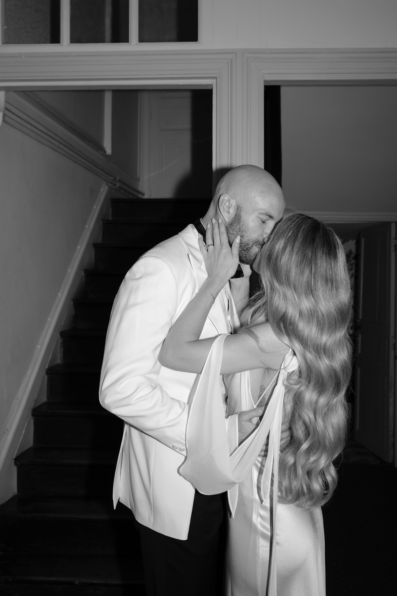 A couple dressed in formal attire shares a kiss in front of a staircase indoors.