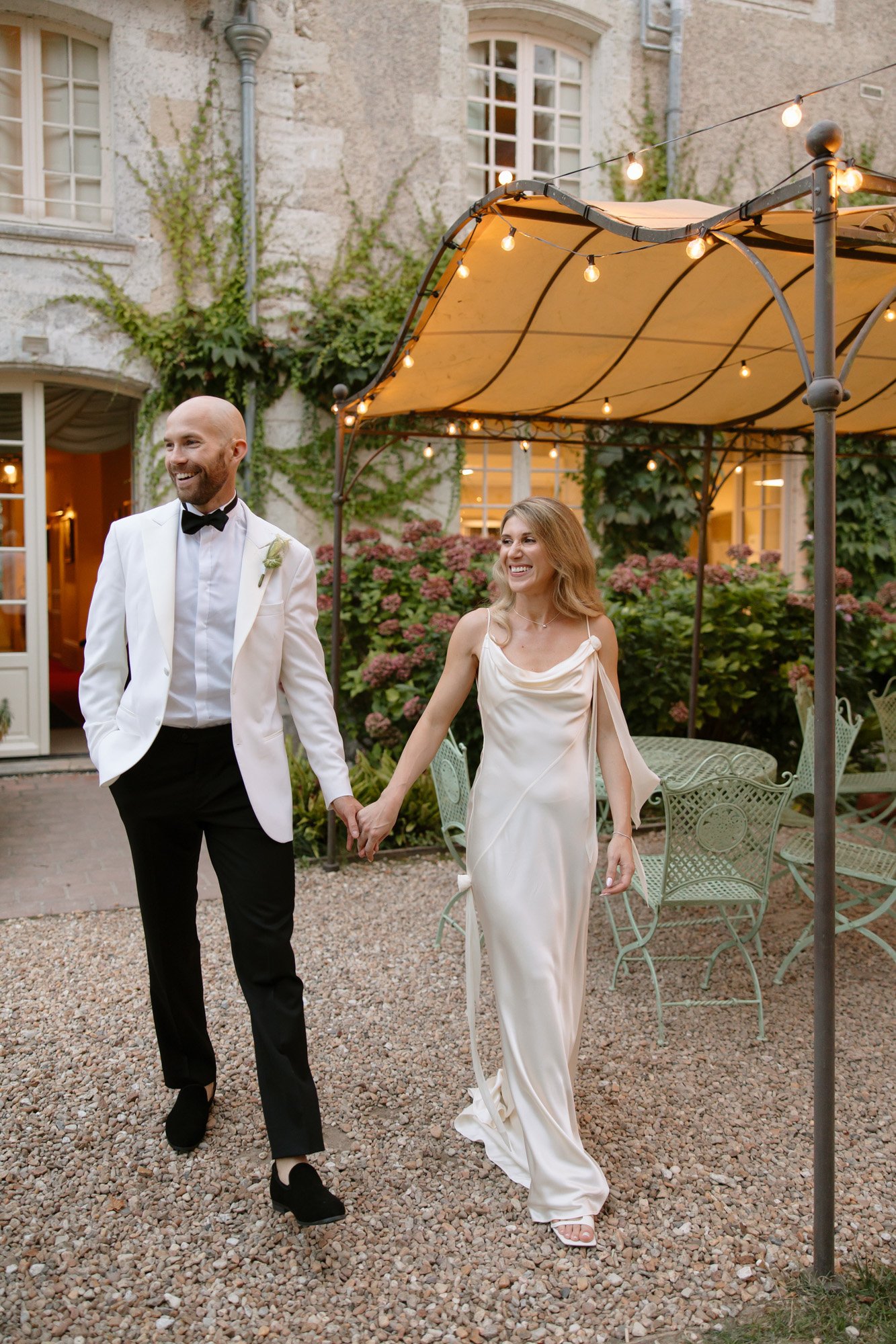 A couple dressed in formal wedding attire hold hands and walk outside under a canopy with string lights, smiling.