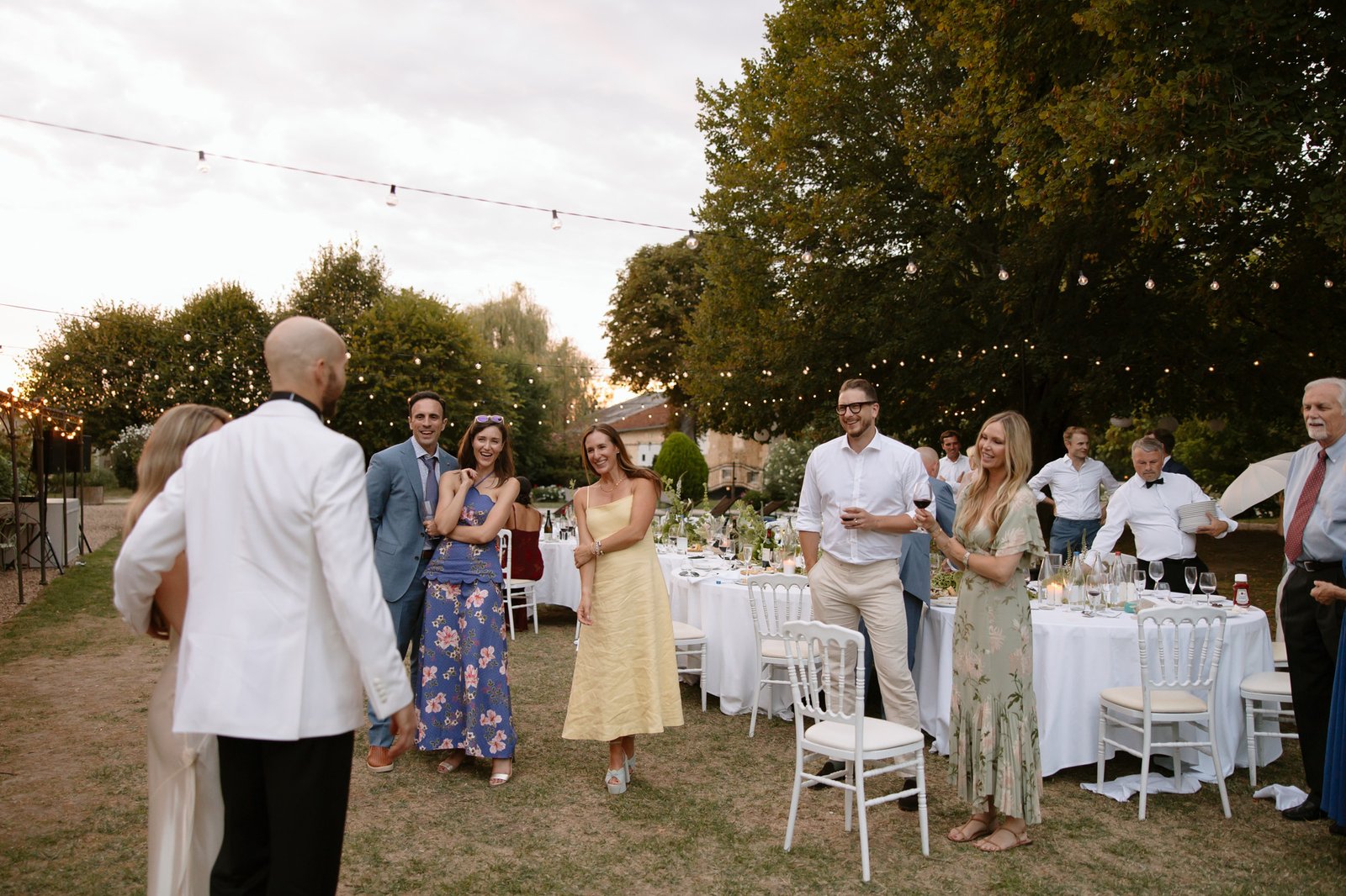 A group of people dressed in semi-formal attire stand and interact outdoors near decorated tables at a garden event during sunset.