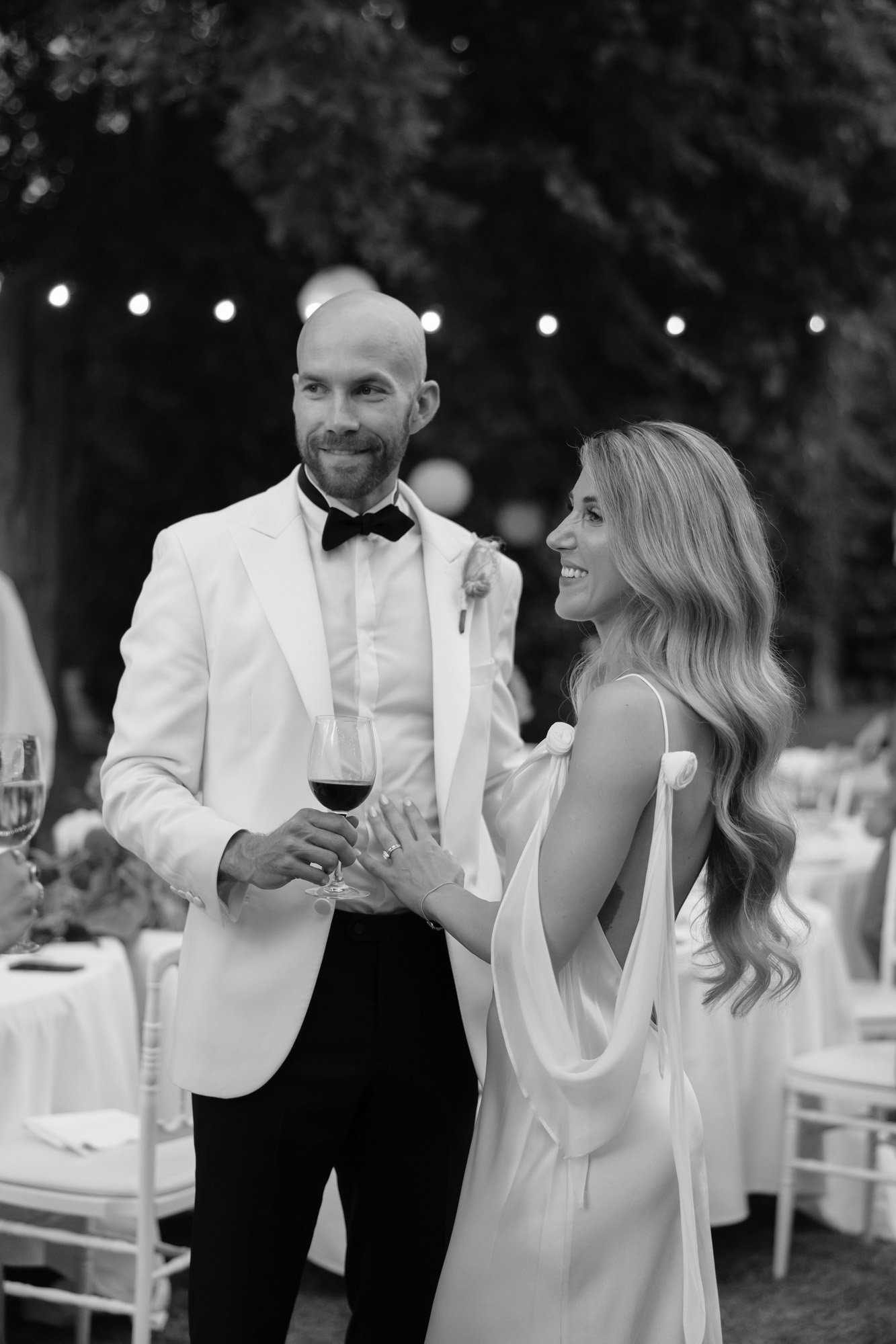 A man in a white tuxedo and a woman in a white gown stand together outdoors, holding wine glasses and smiling, with tables and string lights in the background.