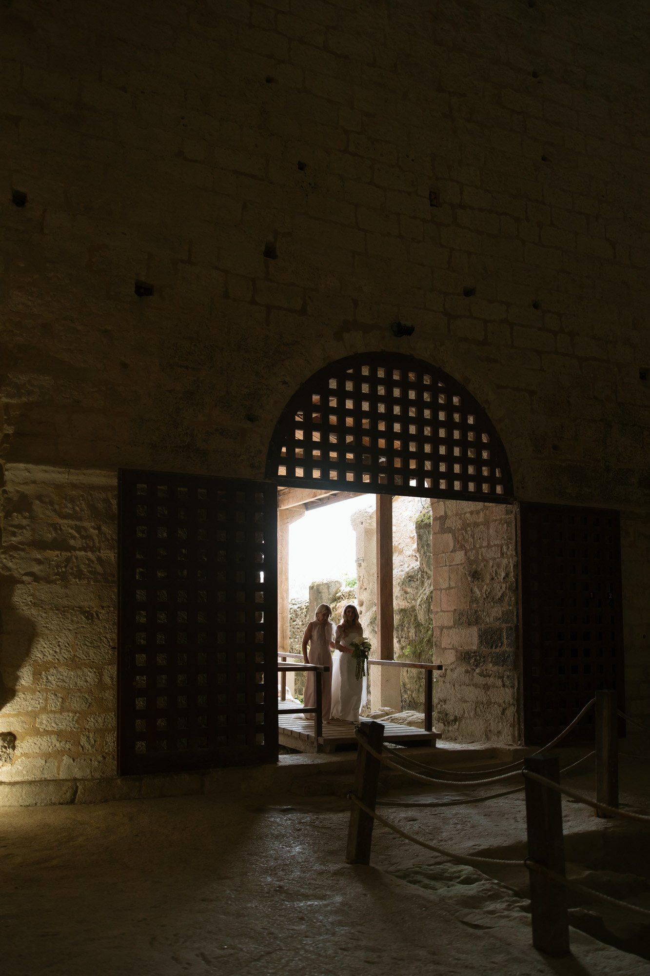 Two people stand near a large arched doorway with a lattice design, illuminated by natural light from outside, inside a stone-walled historic building. Aubeterre sur dronne wedding.
