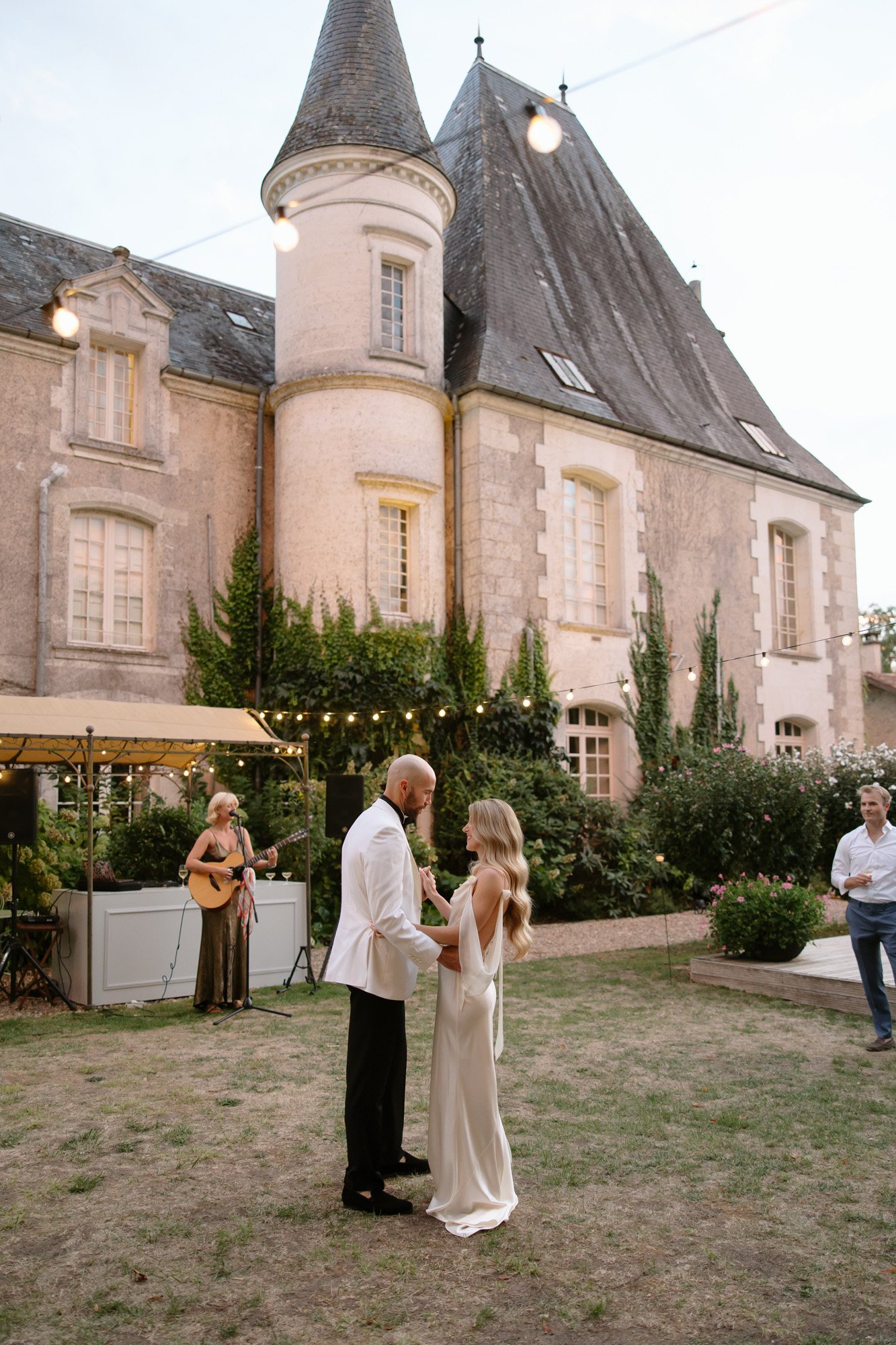 A bride and groom dance outside a stone building with a conical tower, as a musician plays guitar nearby and guests watch.