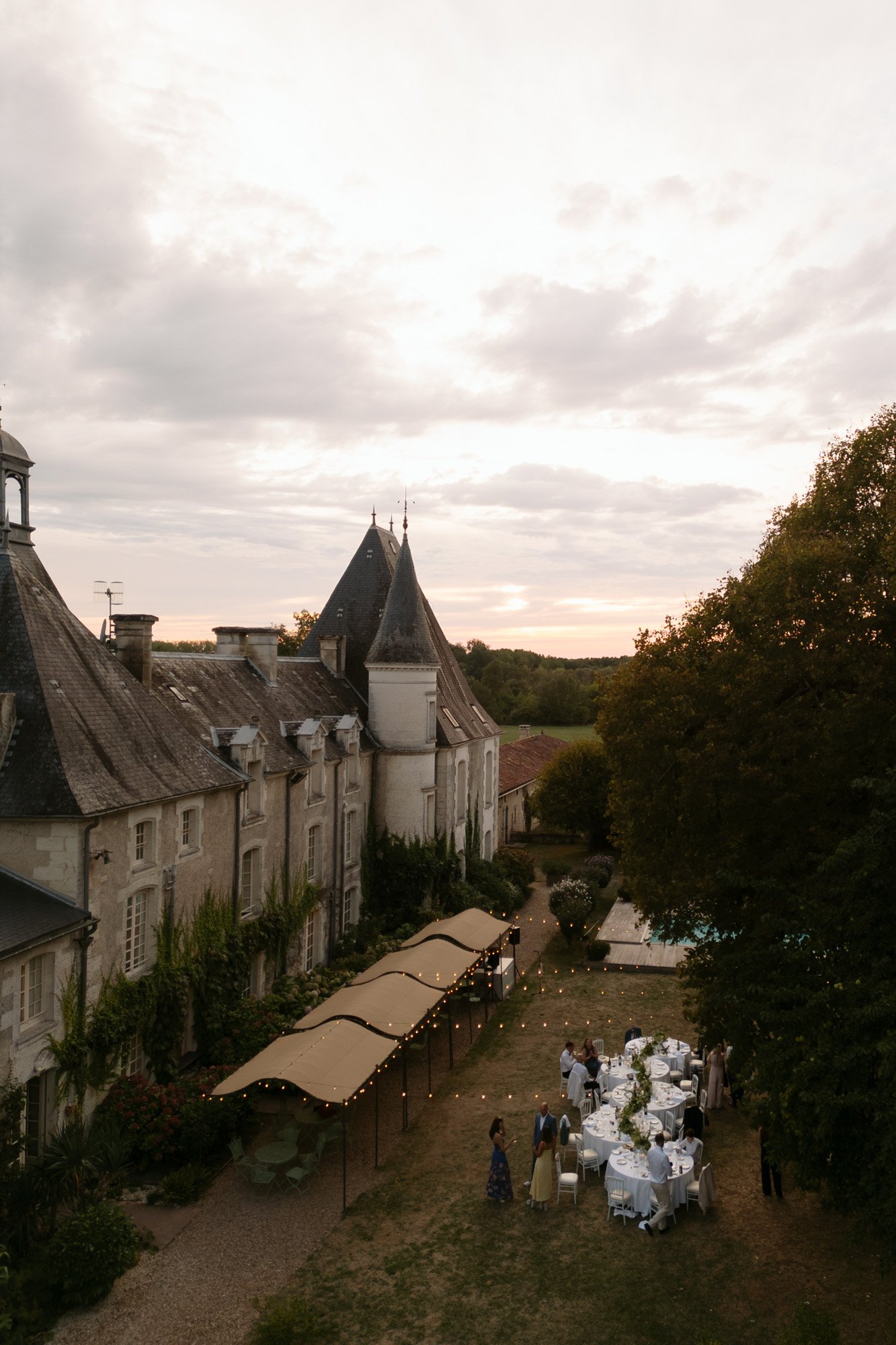 An outdoor dinner gathering with white tablecloths is set up beside a historic stone building with turrets and greenery at sunset.