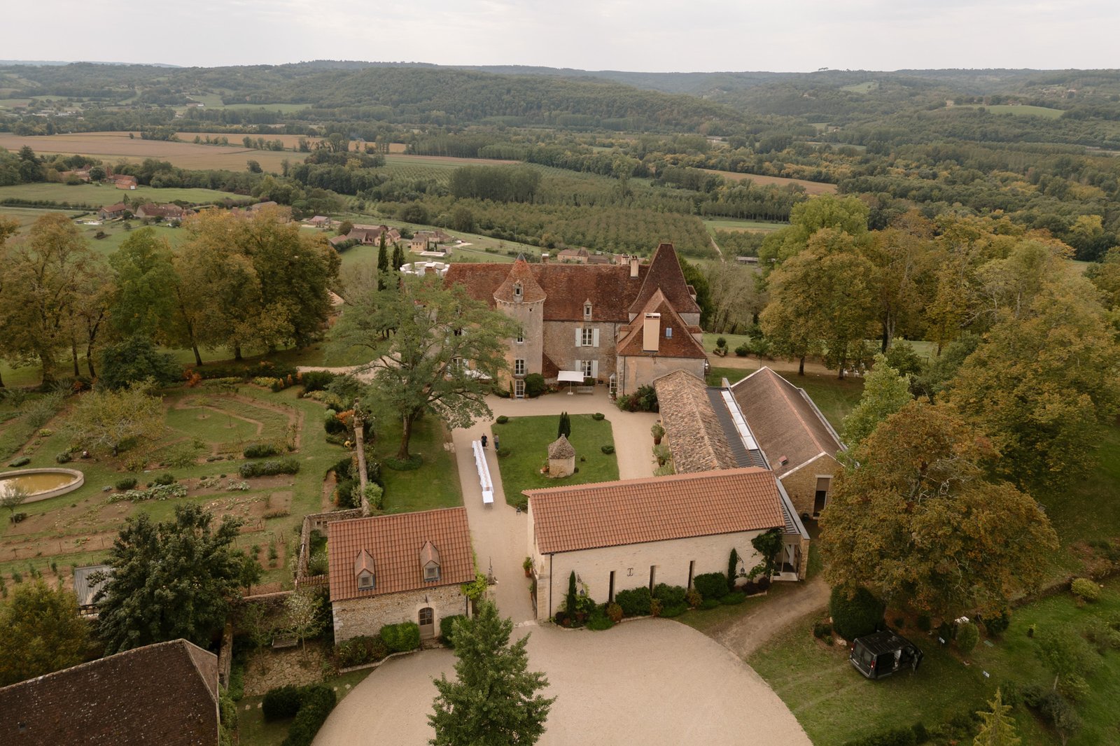 Aerial view of a historic estate with multiple stone buildings, red-tiled roofs, gardens, and surrounding green countryside. Autumn France wedding.