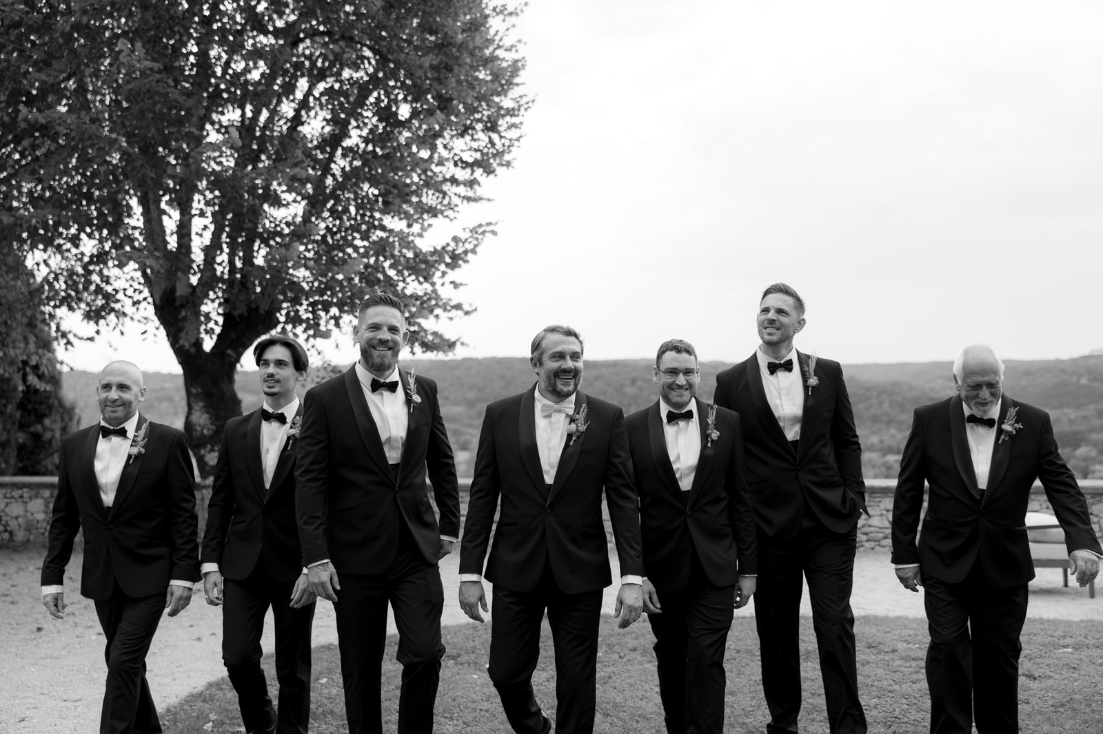 Seven men in black tuxedos and bow ties walk outside together, smiling, with a tree and landscape in the background.