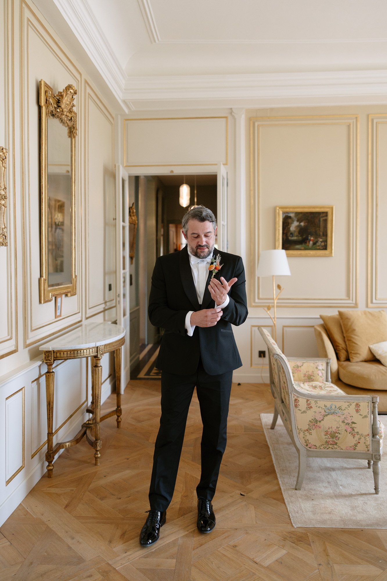 A man in a black tuxedo stands in an elegant room with cream-colored walls, adjusting his cufflink. The room features classic decor, paintings, and ornate furniture. Autumn France wedding.