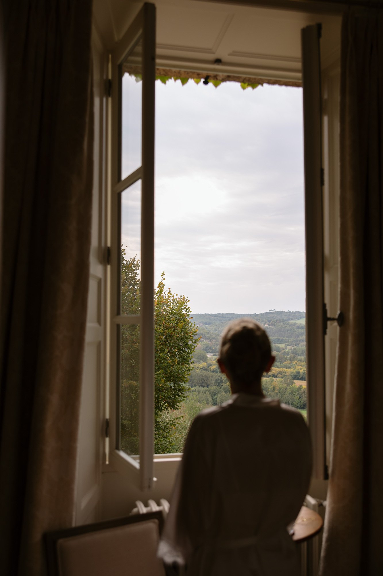 A person stands by an open window, gazing out at a landscape of trees and hills under a cloudy sky.