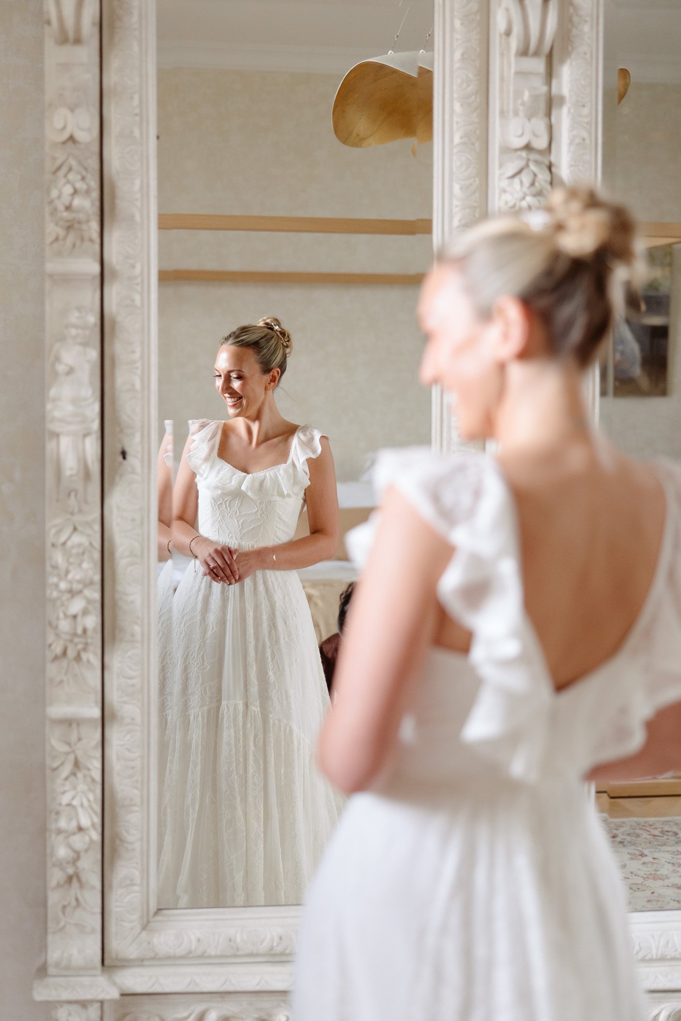 A woman in a white dress with ruffled sleeves stands in front of a large ornate mirror, smiling at her reflection. Autumn France wedding.