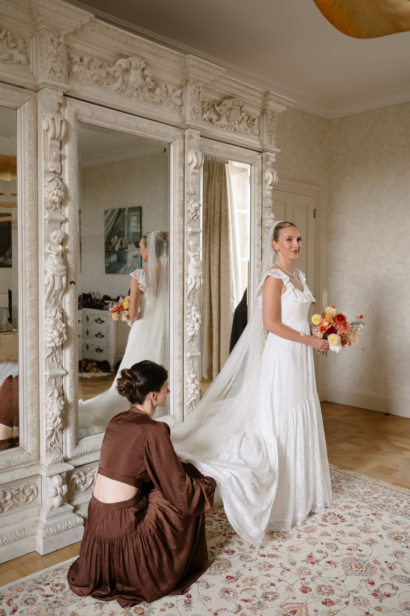 A bride in a white gown stands holding a bouquet while another woman in a brown dress adjusts her train in front of a large ornate mirror. Autumn France wedding.
