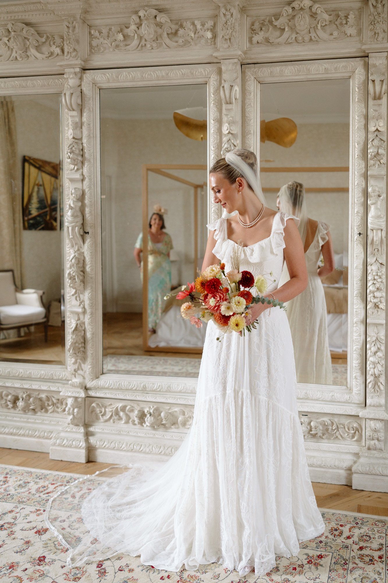 A bride in a white dress holding a bouquet of flowers stands in front of an ornate mirrored wall, with another woman reflected in the background. Autumn France wedding.