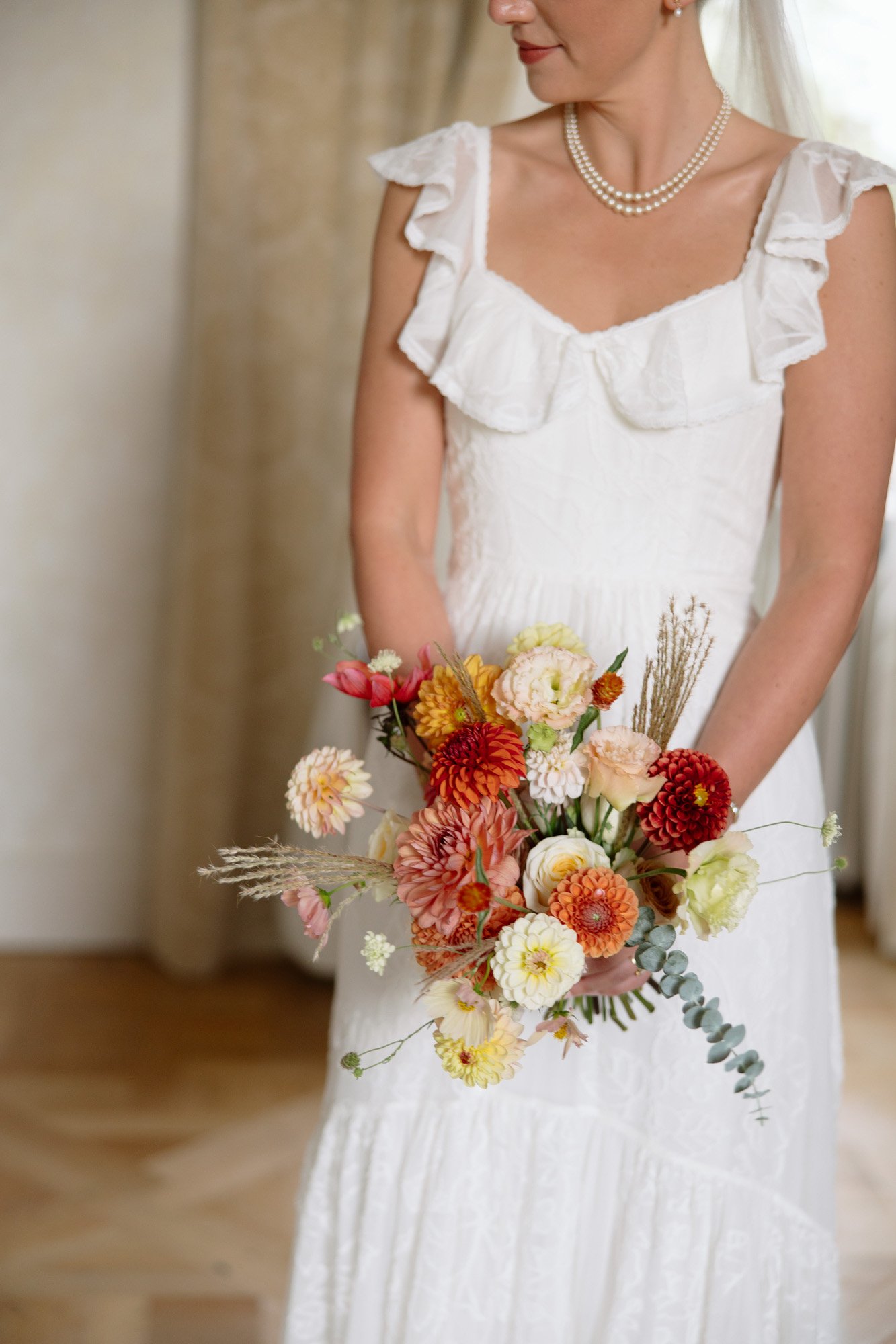 A woman in a white dress with ruffled sleeves holds a colorful bouquet of flowers. She wears a pearl necklace and veil. Autumn France wedding.