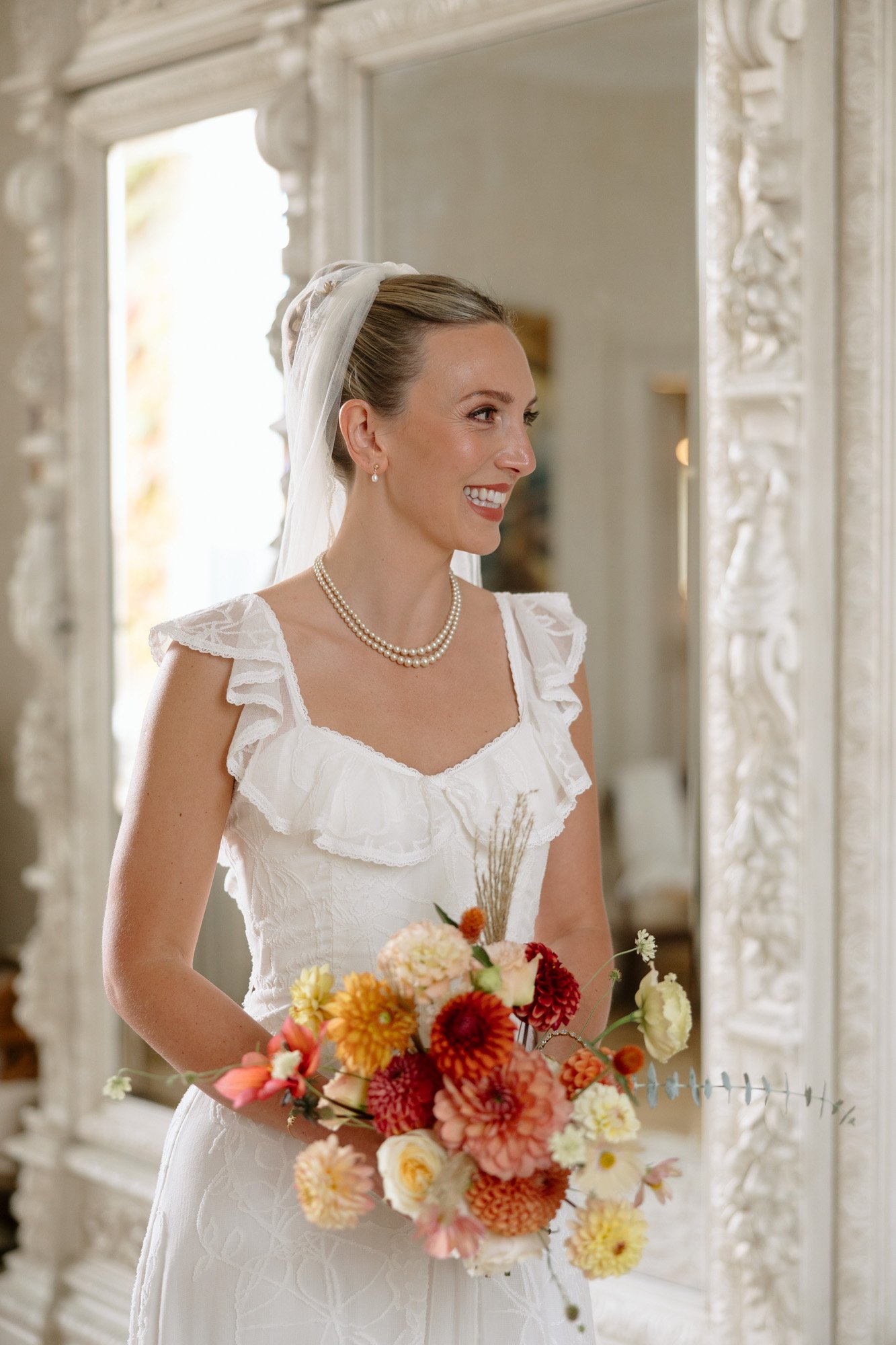 A woman in a white bridal dress and veil holds a bouquet of colorful flowers, standing in front of an ornate white mirror and smiling. Autumn France wedding.