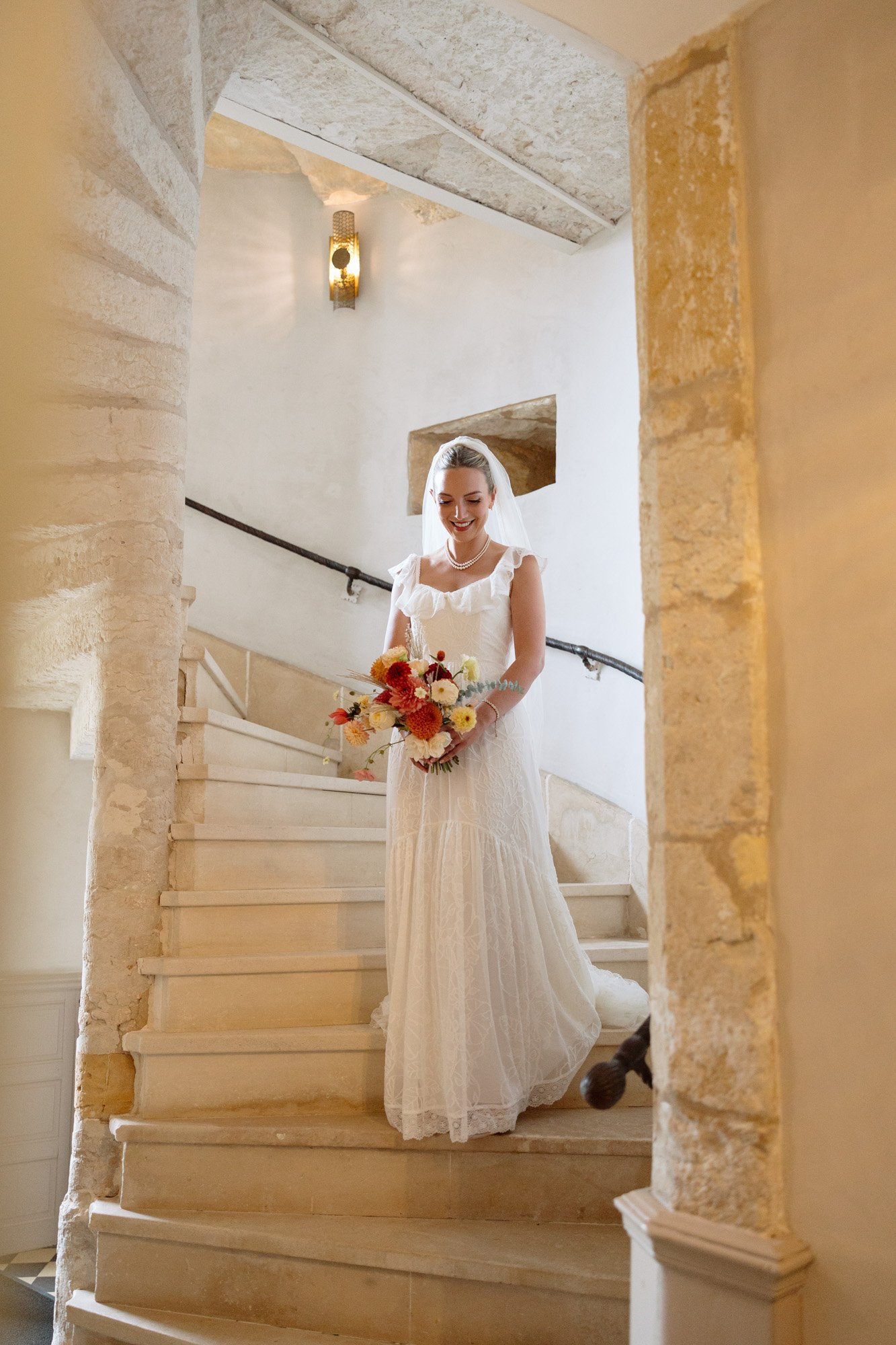A bride in a white dress and veil holds a bouquet of flowers while walking down a stone staircase in a well-lit interior. Autumn France wedding.