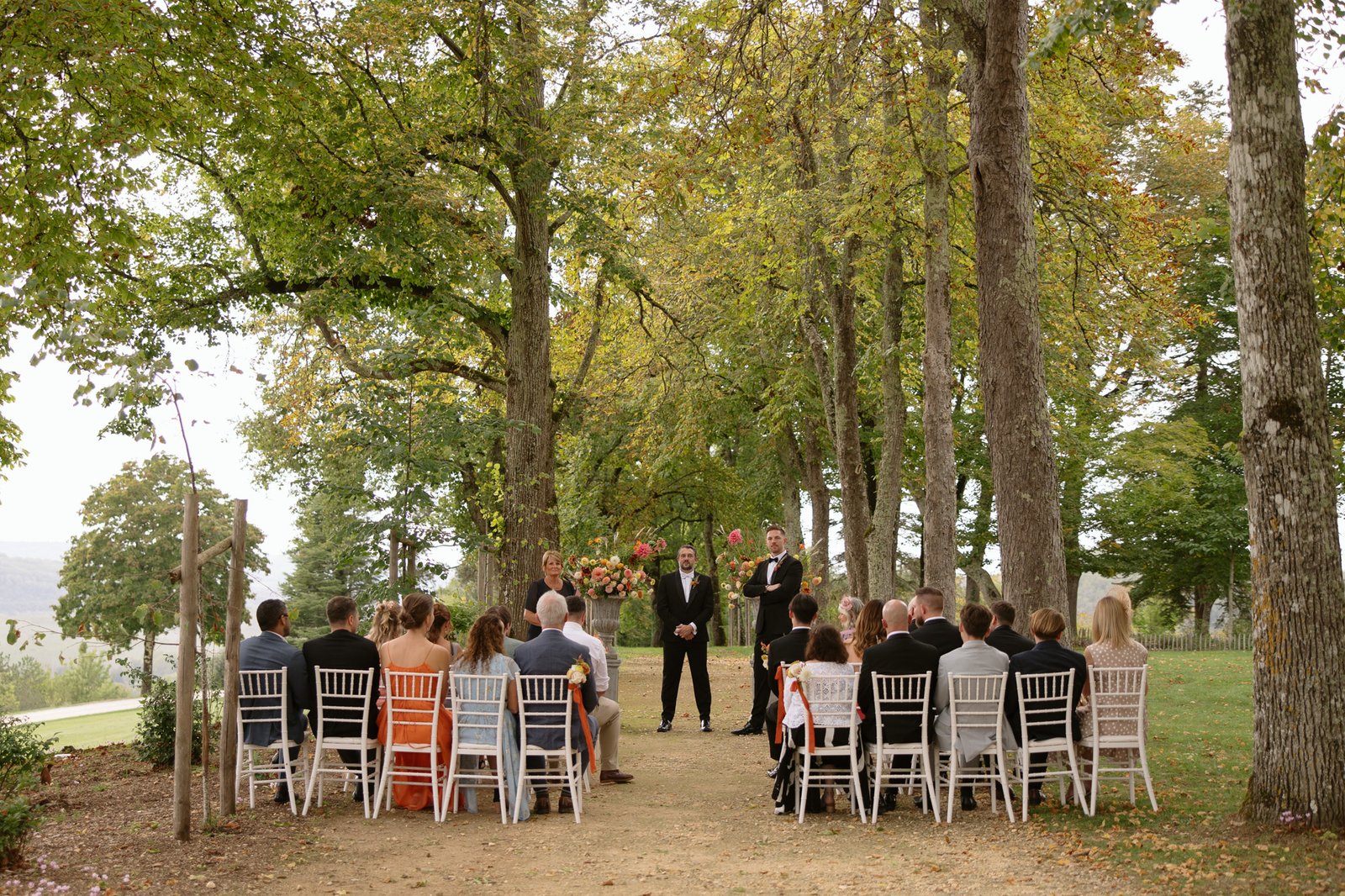 Outdoor wedding ceremony with guests seated on white chairs facing an officiant, groom, and best man under tall trees on a dirt path. Autumn France wedding.