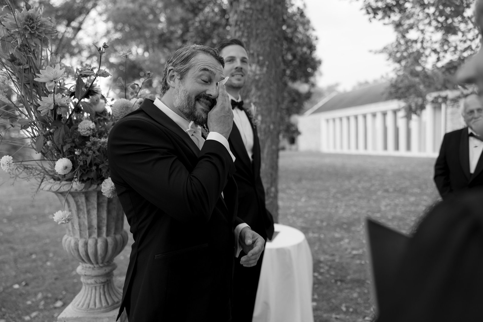 A man in a suit wipes away a tear while standing outdoors next to a flower arrangement, with other men in suits nearby.