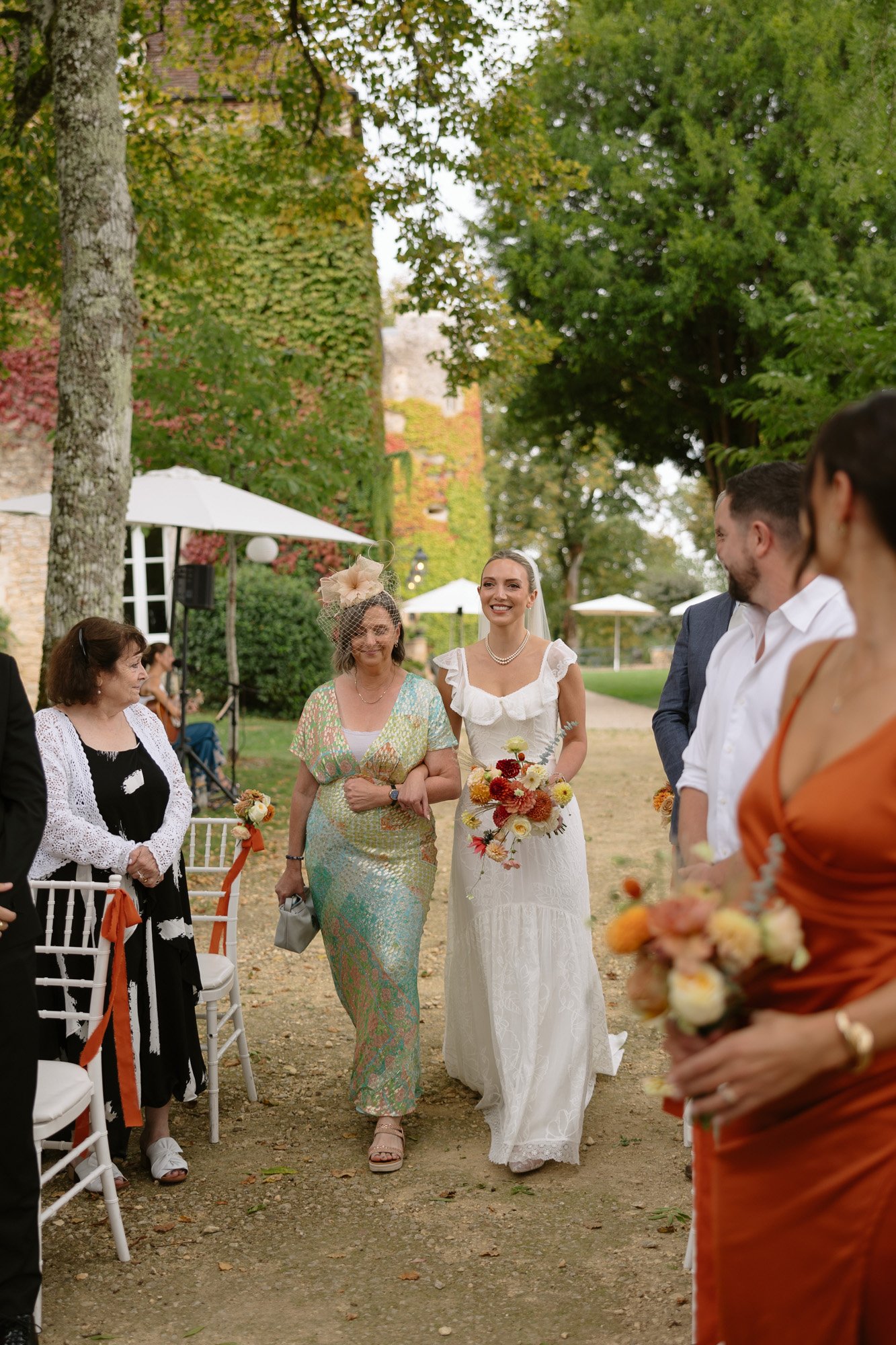 A bride in a white dress walks down an outdoor aisle with a woman in a light blue and gold dress, surrounded by seated and standing guests. Autumn France wedding.