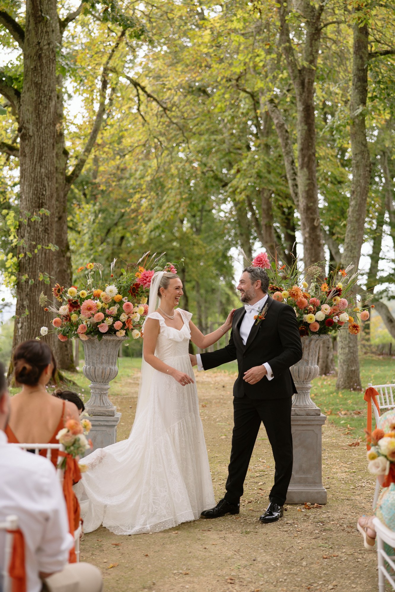 A bride and groom stand smiling together outdoors, dressed in wedding attire, with floral arrangements and guests seated around them. Autumn France wedding.