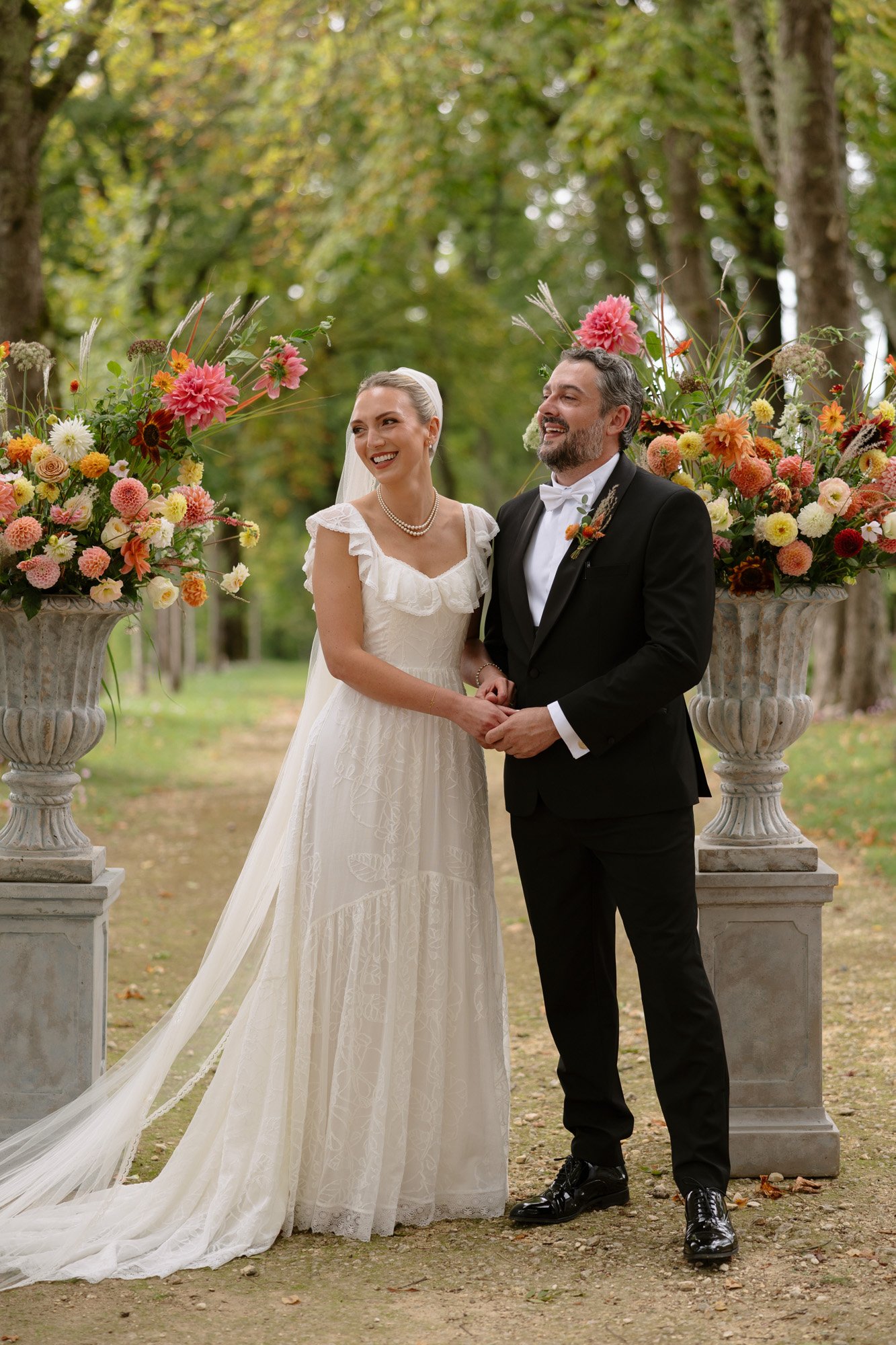 A bride in a white dress and a groom in a black suit stand holding hands and smiling outdoors between two large floral arrangements. Autumn France wedding.