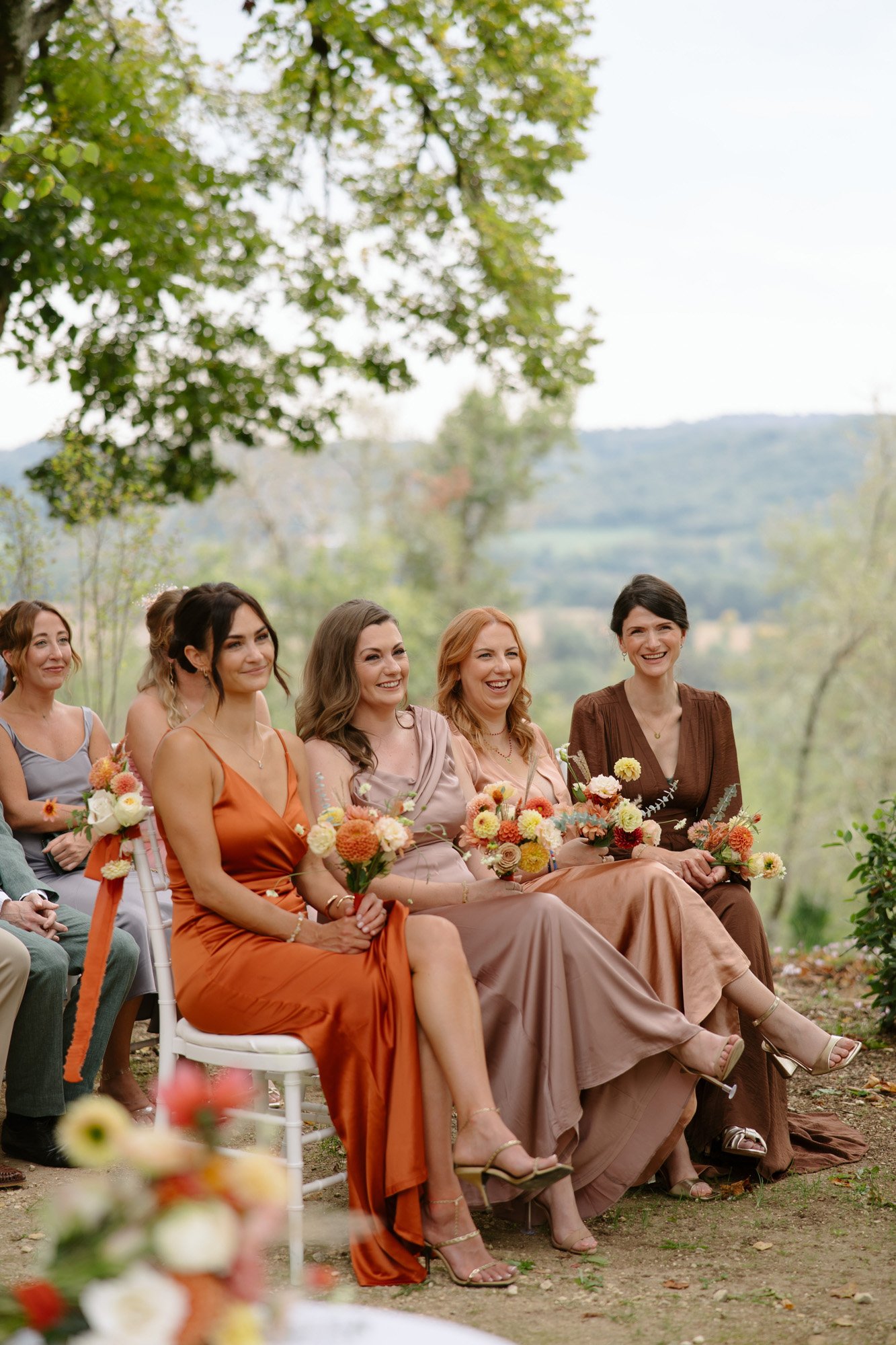 Four women in formal dresses, seated outdoors at an event, holding bouquets and smiling; trees and hills are visible in the background.