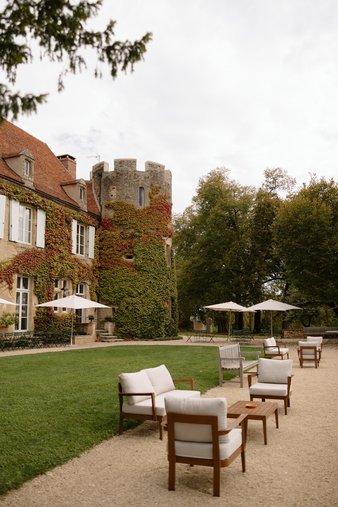 Outdoor seating area with cushioned chairs and umbrellas set on gravel beside a historic ivy-covered stone building with a round tower and red roof, surrounded by trees.