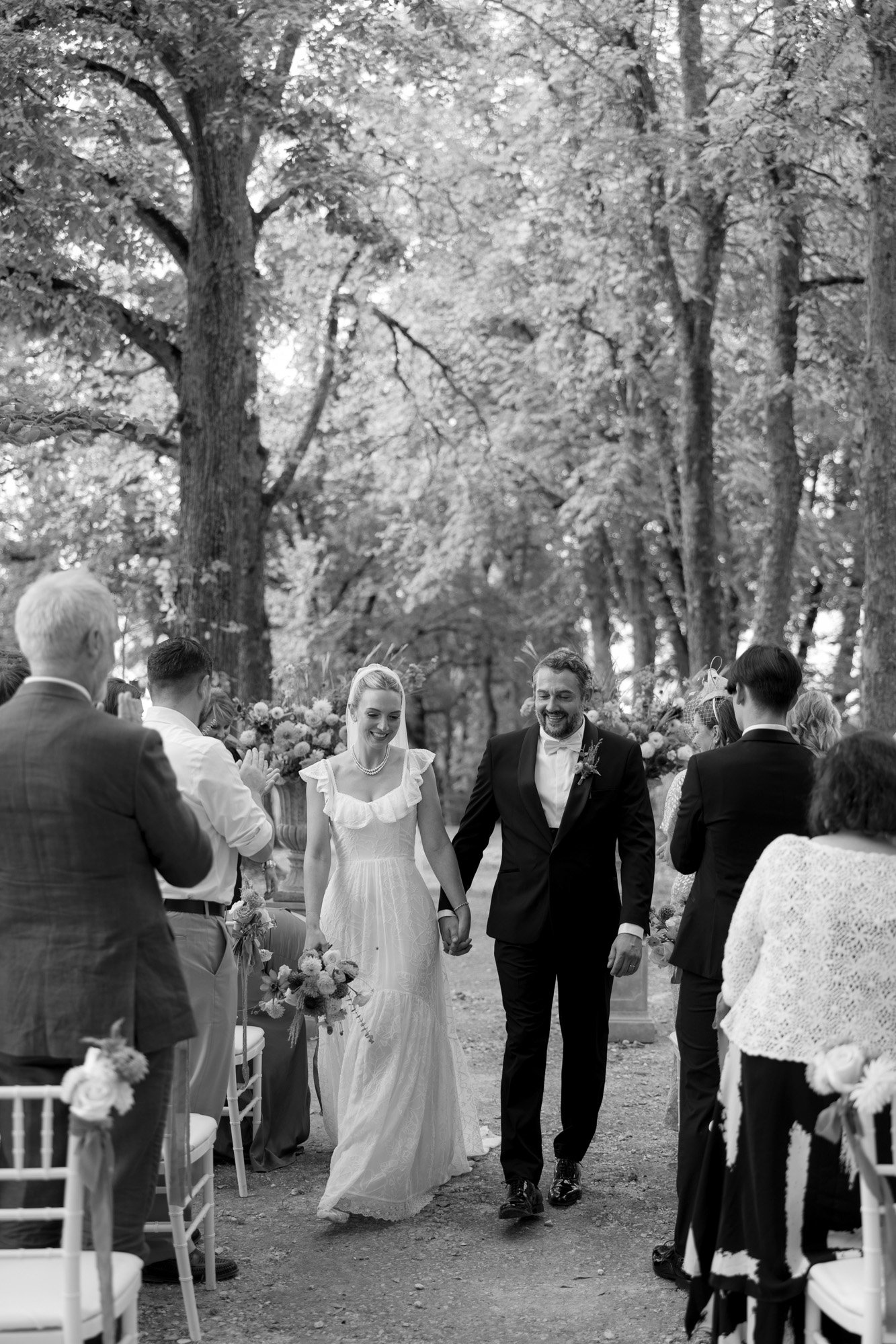 A bride and groom walk hand in hand down an outdoor aisle, surrounded by standing guests and trees, during a wedding ceremony.