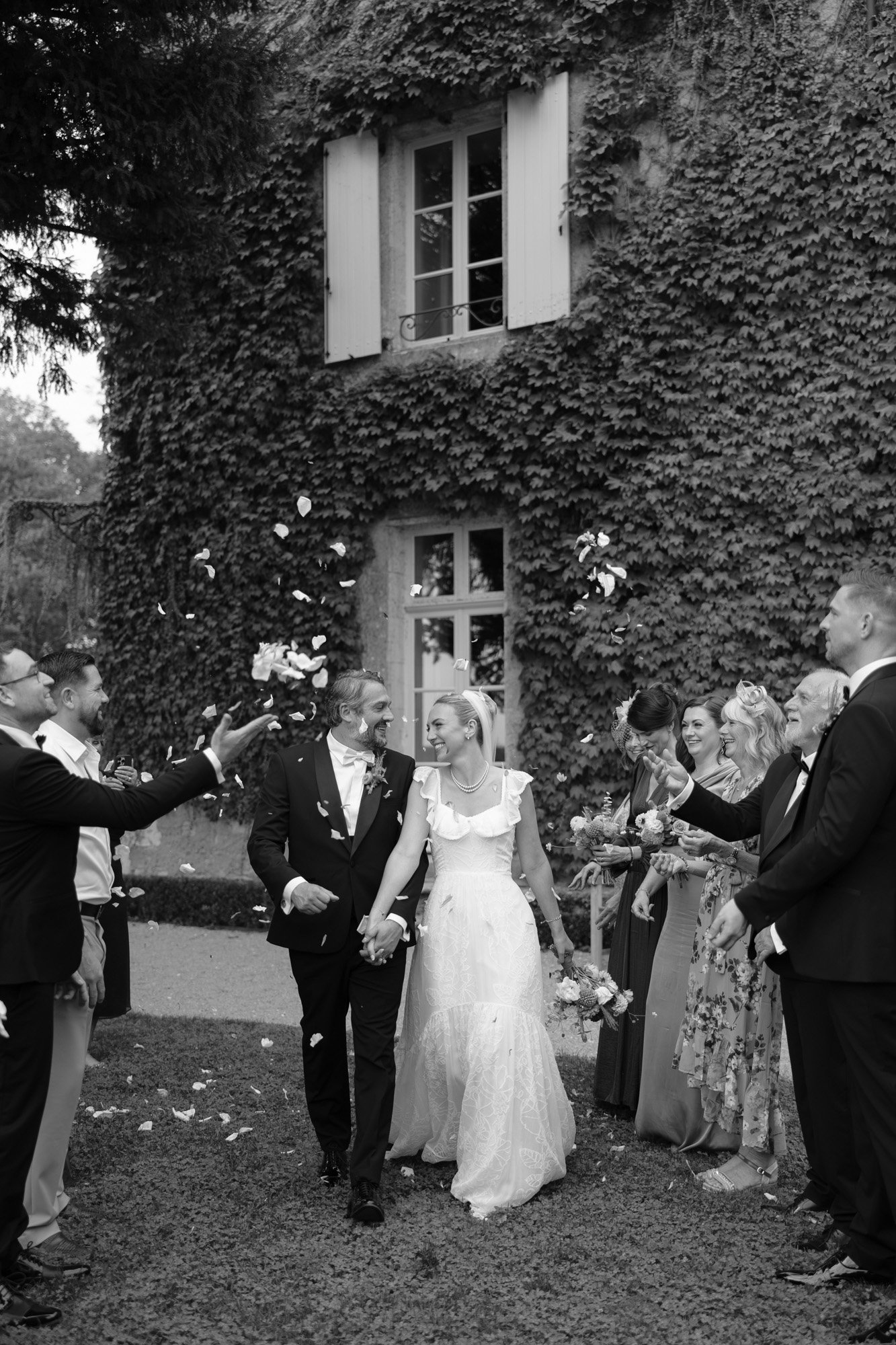 A bride and groom walk hand in hand outside an ivy-covered building while guests throw flower petals and celebrate around them.