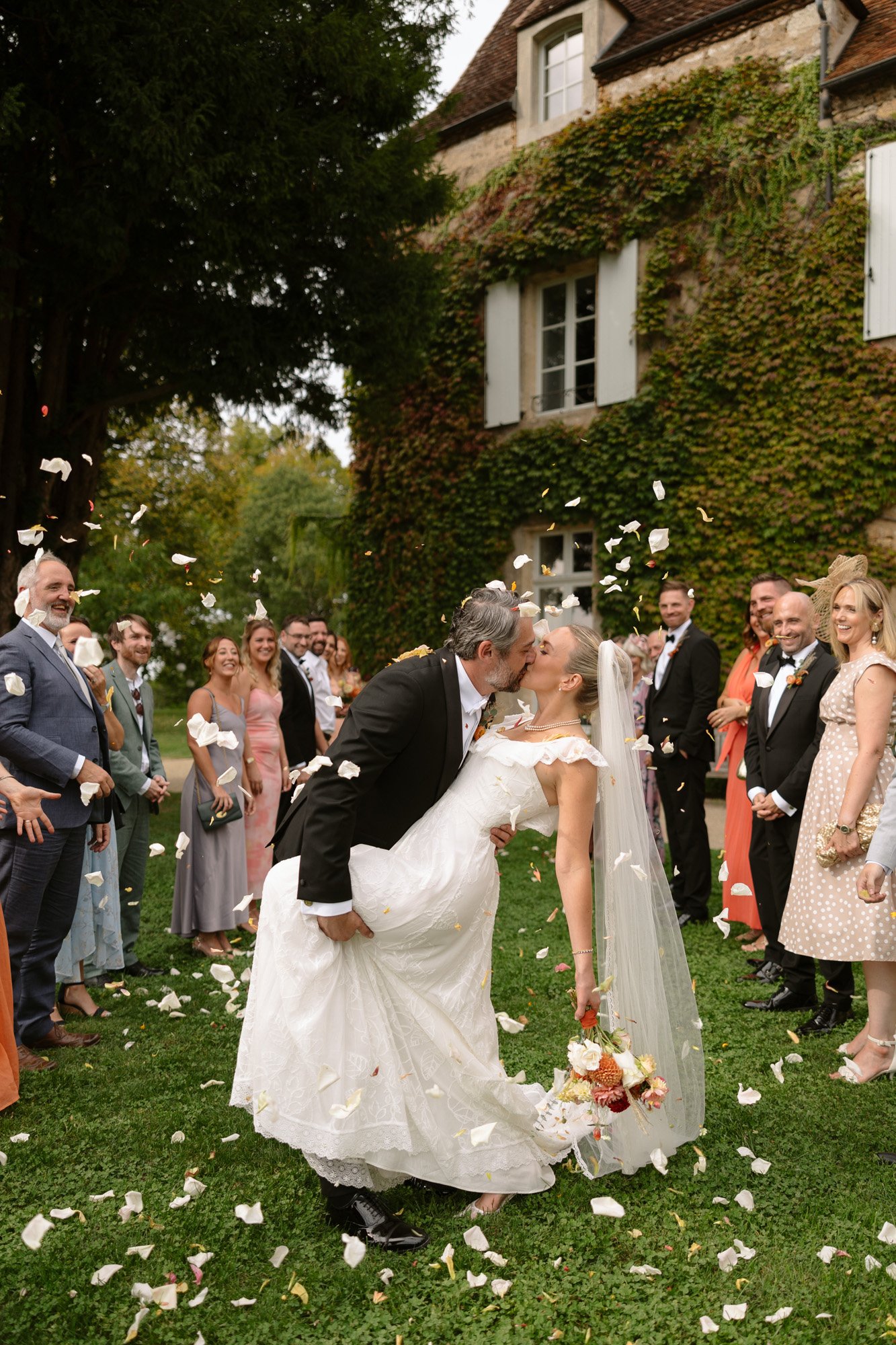 A bride and groom kiss as the groom dips the bride, surrounded by cheering guests outdoors with flower petals in the air. Autumn France wedding.