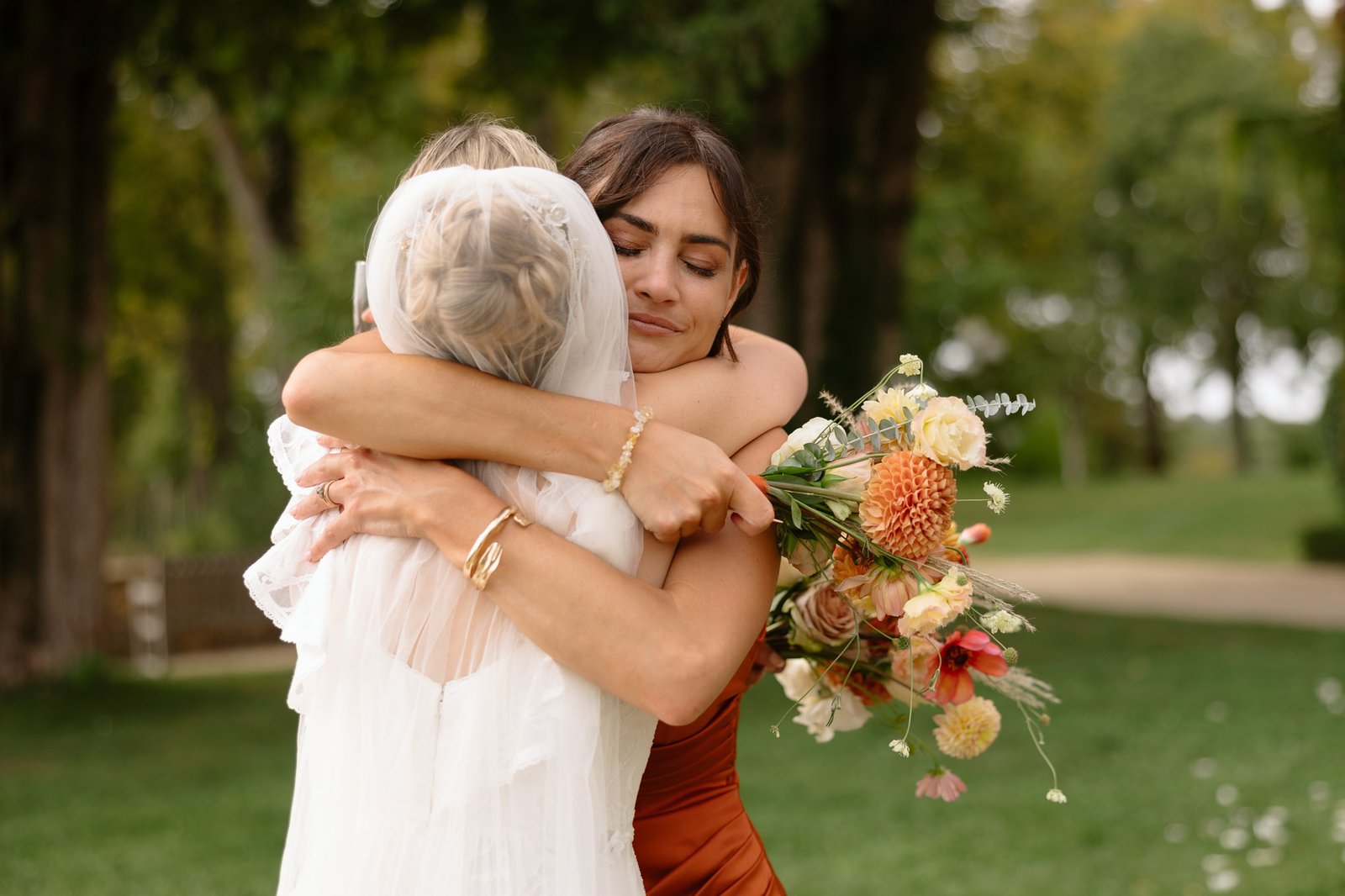 Two women embrace outdoors; one wears a white wedding dress and veil, holding a bouquet of flowers, while the other wears a brown dress. Trees and grass appear in the background. Autumn France wedding.