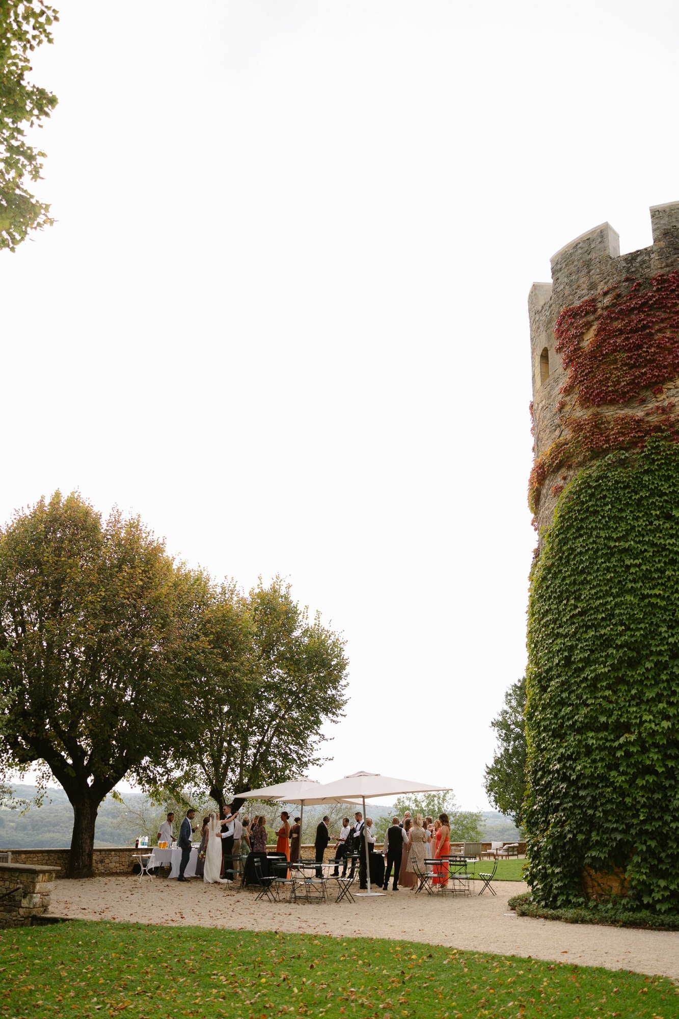 A group of people gathers under a white canopy near a stone tower covered in green ivy, with trees and outdoor seating in the background. Autumn France wedding.
