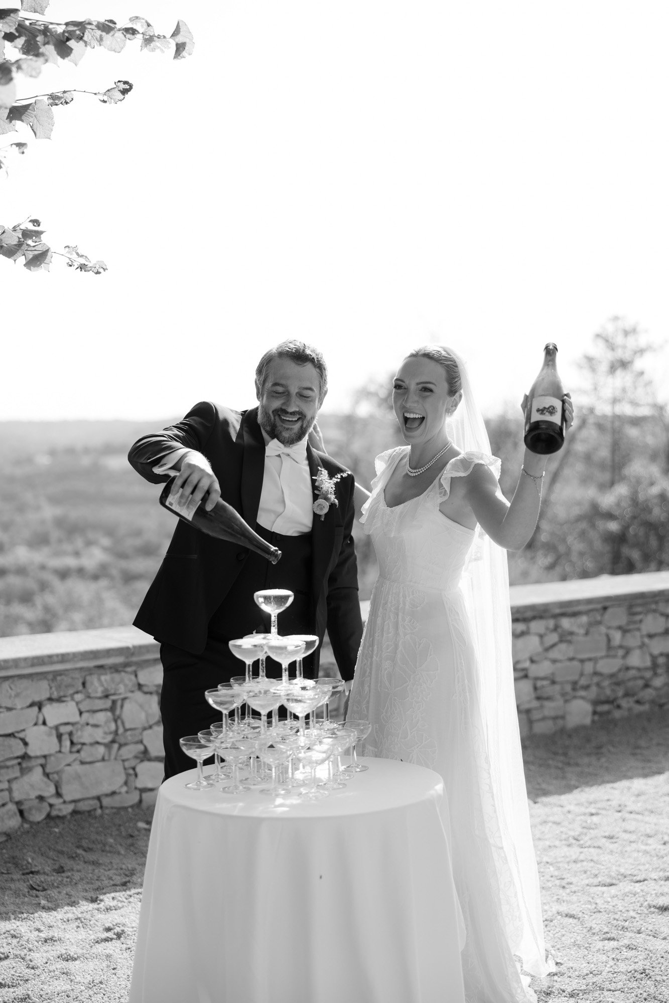 A bride and groom pour champagne into a tower of glasses outdoors, smiling and celebrating in formal wedding attire. Autumn France wedding.