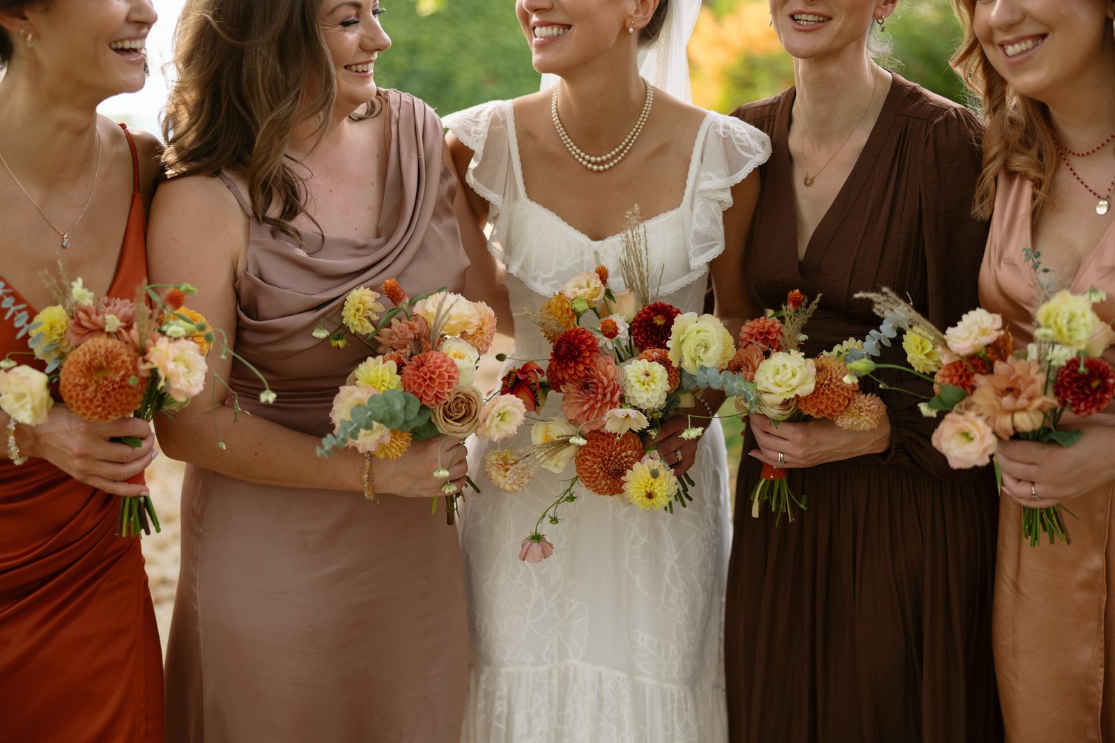 Five women dressed in formal gowns, including a bride in white, stand together holding floral bouquets and smiling. Autumn France wedding.