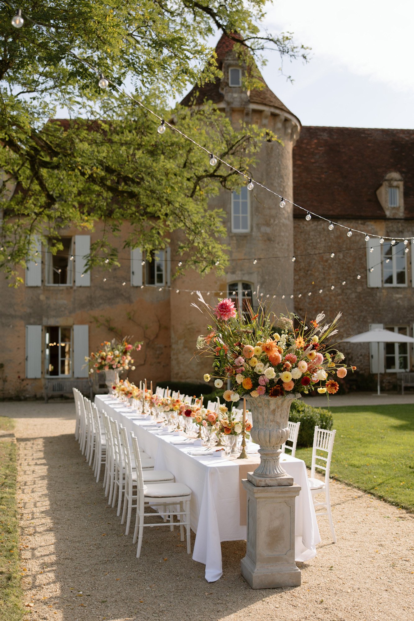 A long outdoor dining table with white chairs is set with floral centerpieces and tableware, situated beside a historic stone building under string lights and a tree. Autumn France wedding.