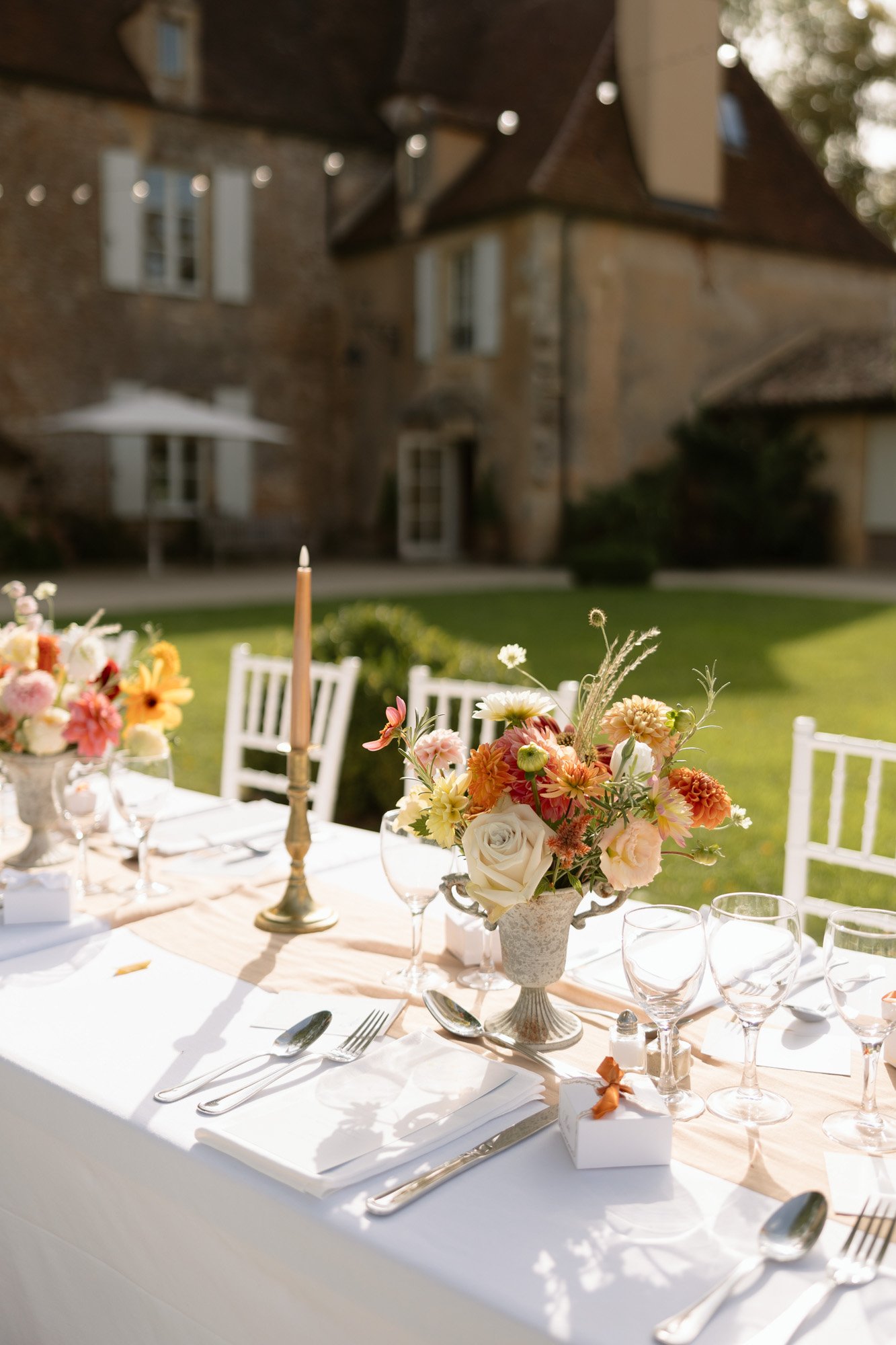 A white table set for an outdoor event with floral centerpieces, glassware, cutlery, and a tall candle in front of a stone building. Autumn France wedding.