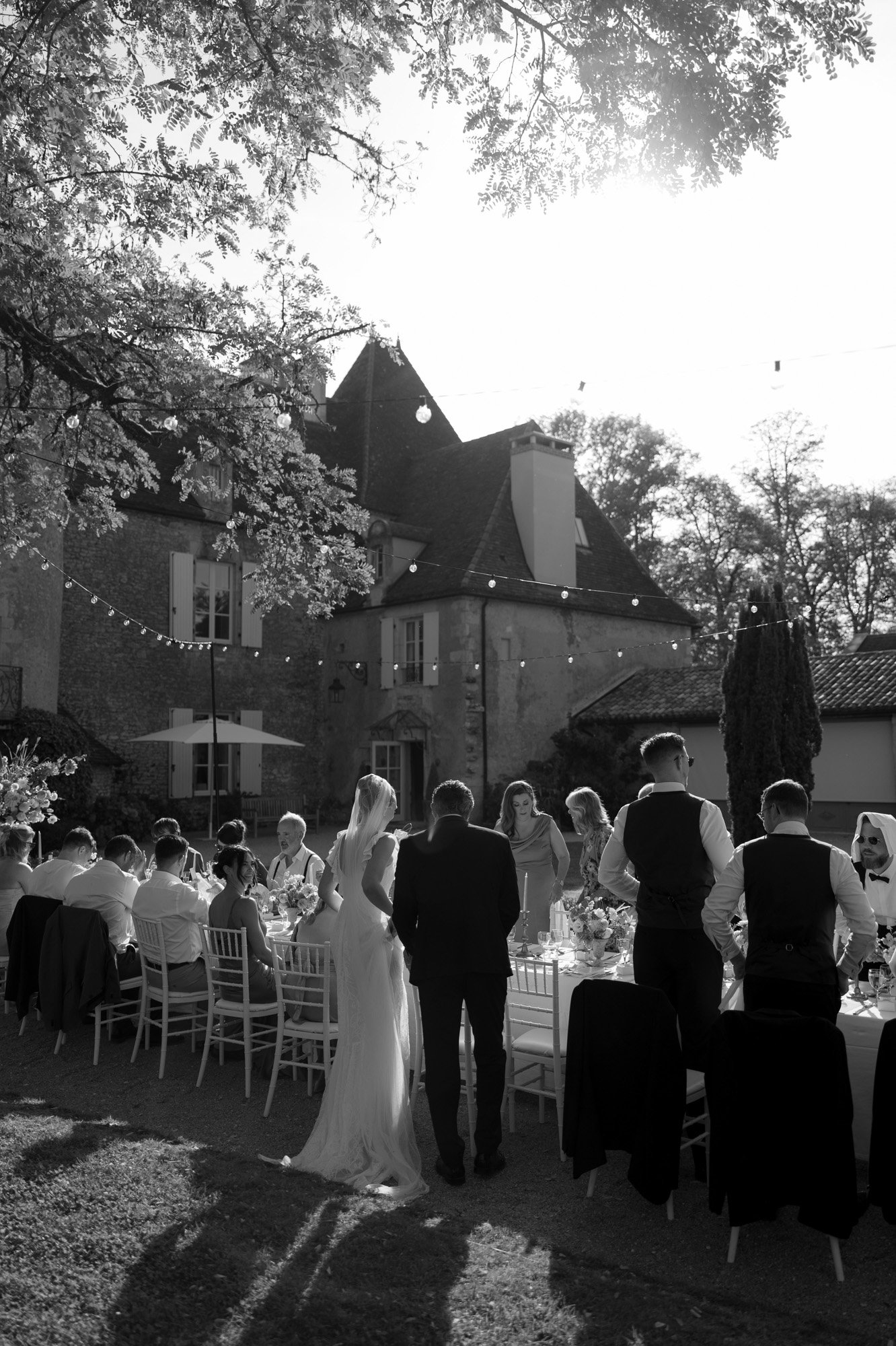 A wedding reception held outdoors beside a large house, with guests seated at long tables and the bride and groom standing together in the foreground.