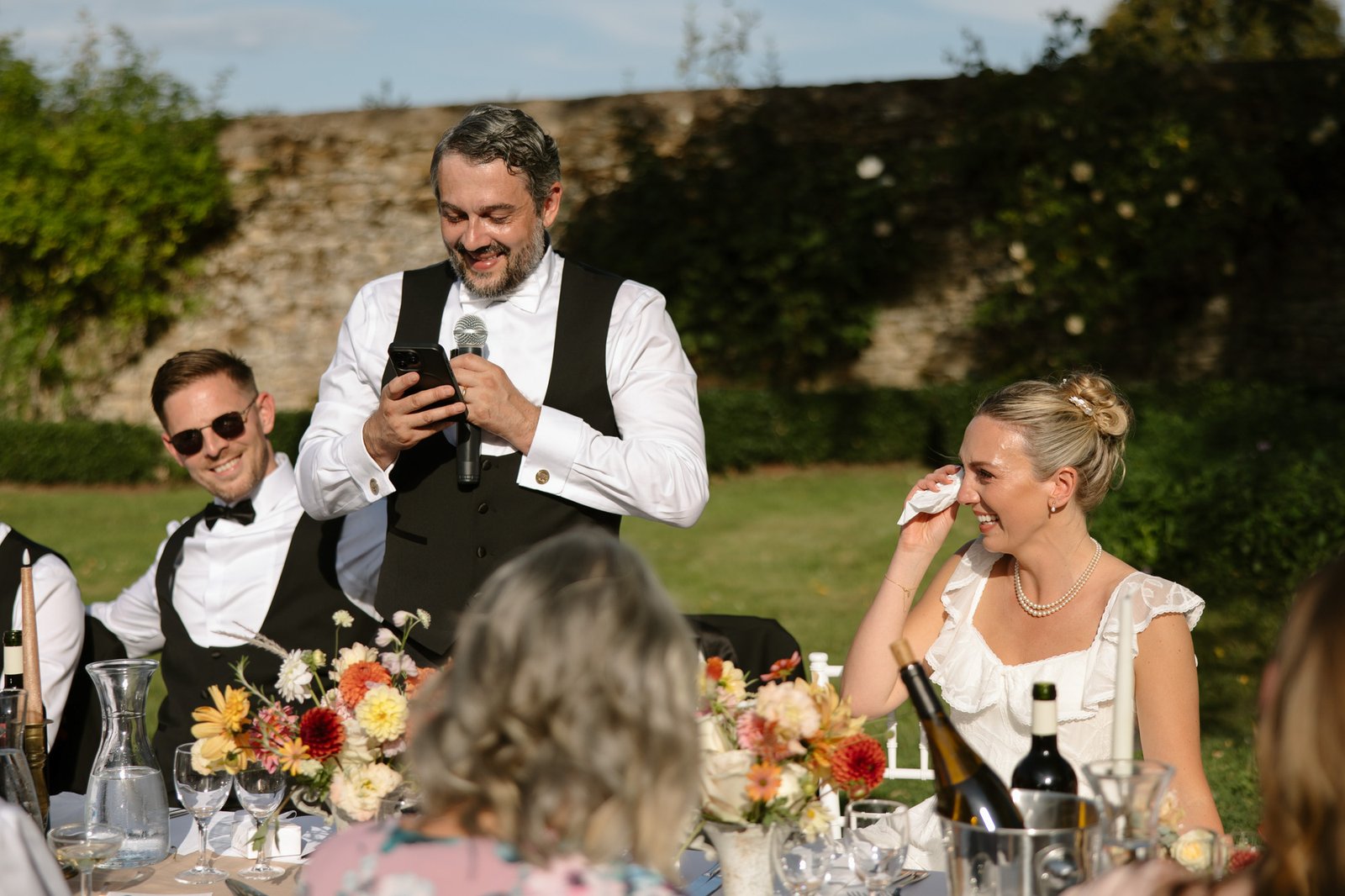 A man in a vest reads from his phone into a microphone at an outdoor table; a woman beside him smiles and wipes her eye, guests and floral arrangements in view. Autumn France wedding.