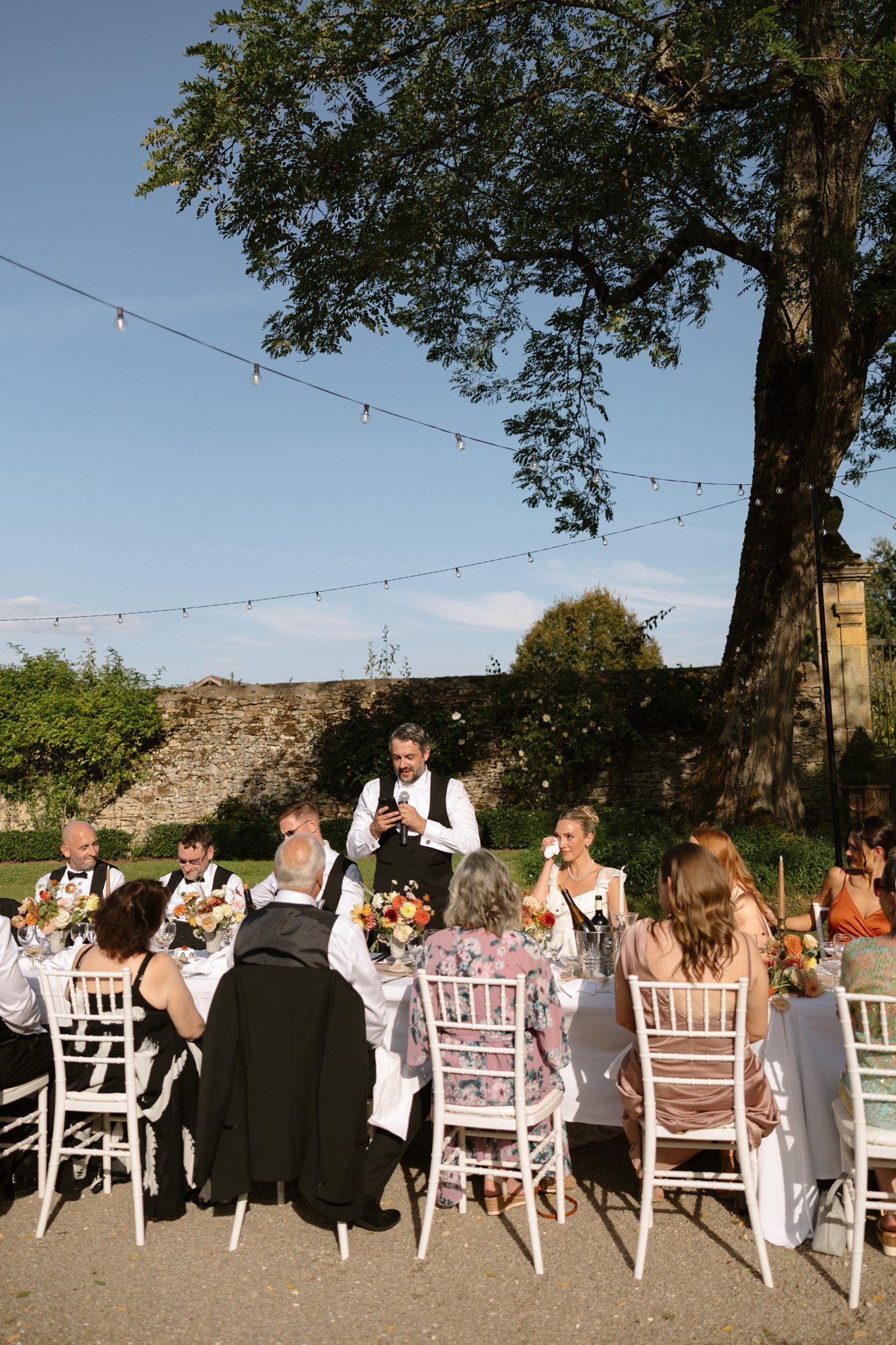 A man in a vest stands giving a speech at an outdoor event, with guests seated around a long table decorated with flowers under string lights. Autumn France wedding.
