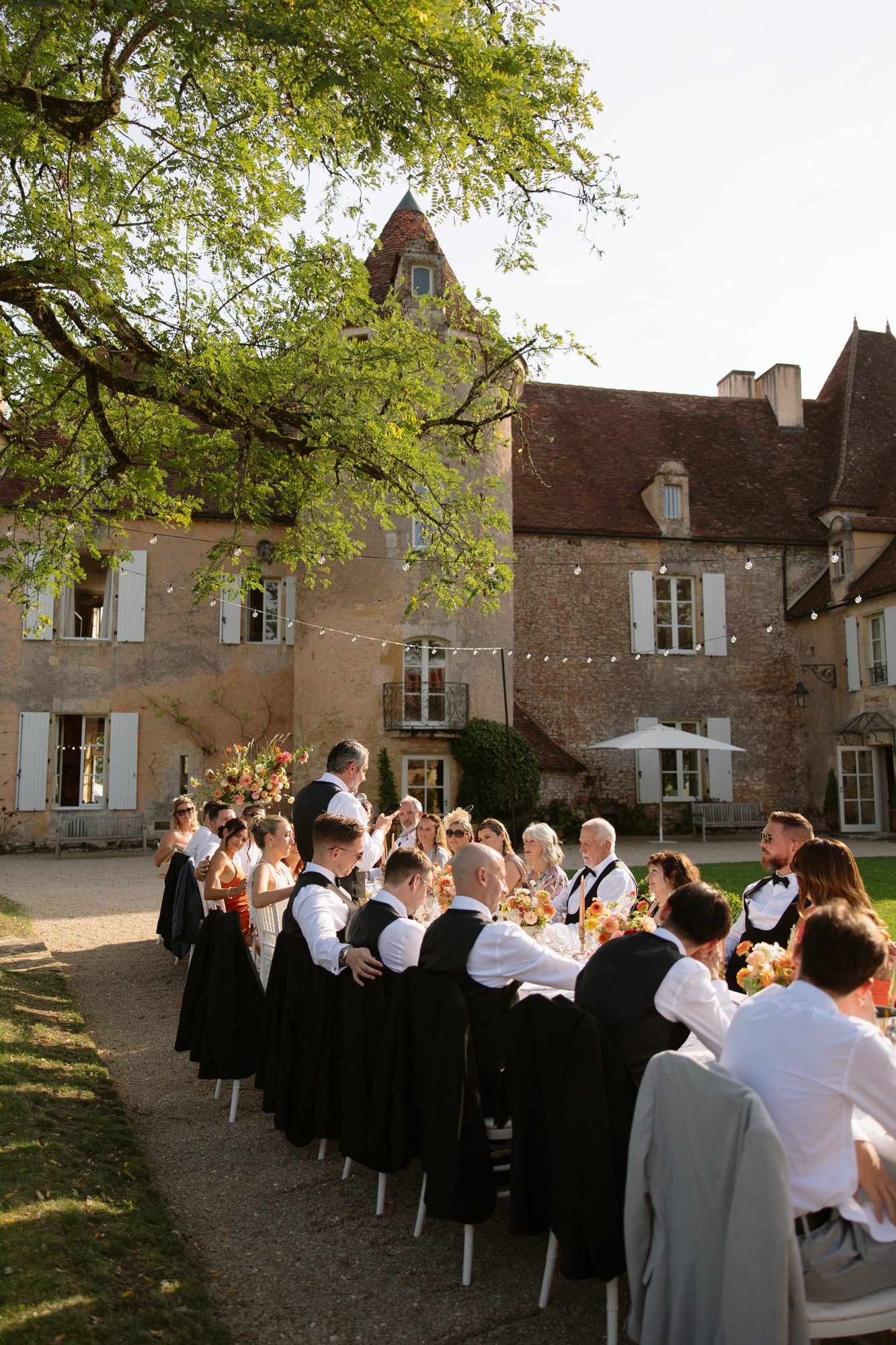 A group of people dressed formally are seated at a long outdoor table for a meal in front of a large, old stone building on a sunny day. Autumn France wedding.