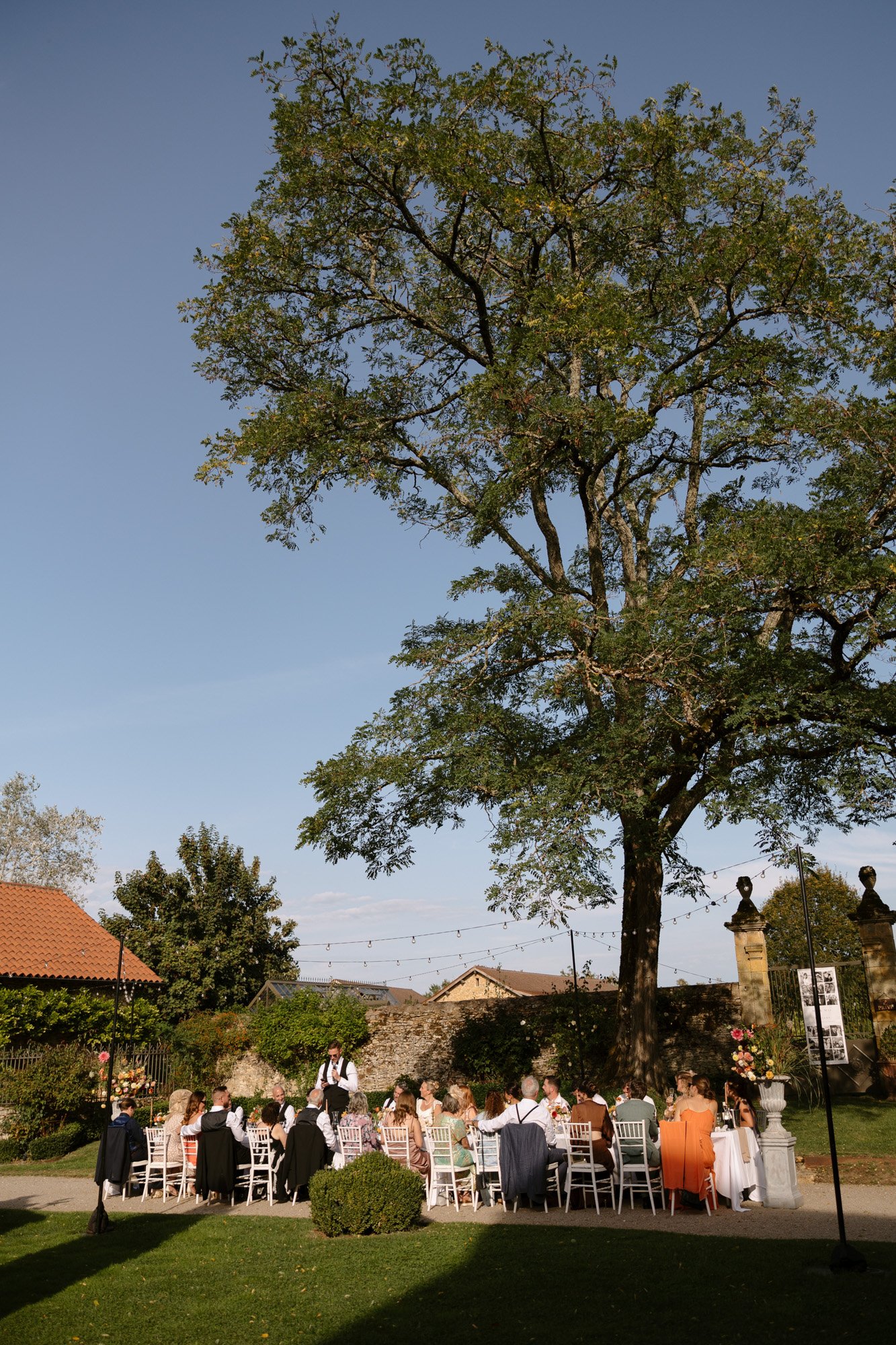 A group of people are seated at tables outdoors under a large tree, attending a formal event on a sunny day.
