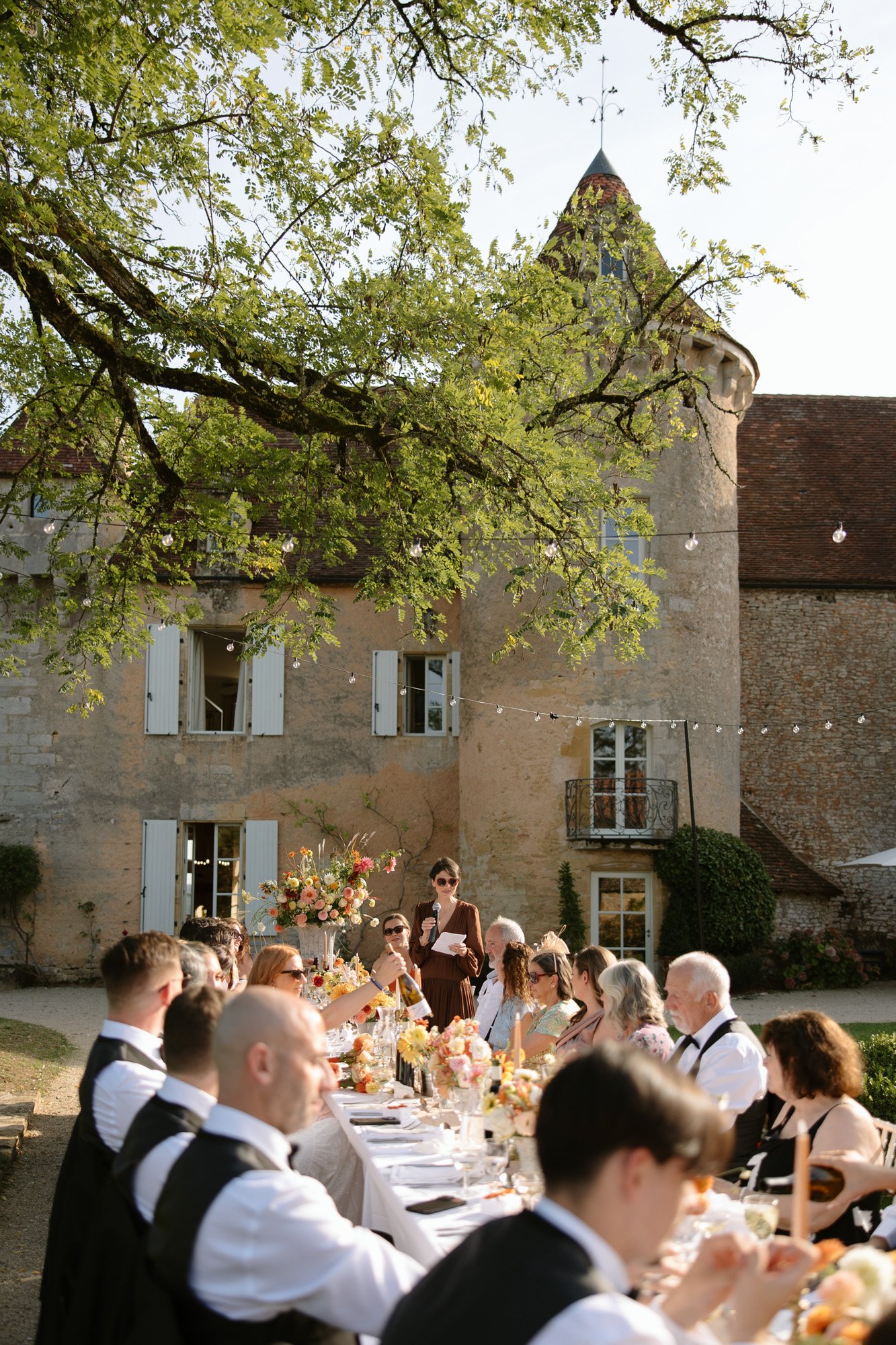 A group of people sit at a long outdoor table set for a meal in front of a stone building with white shutters, while one person stands and speaks. Autumn France wedding.