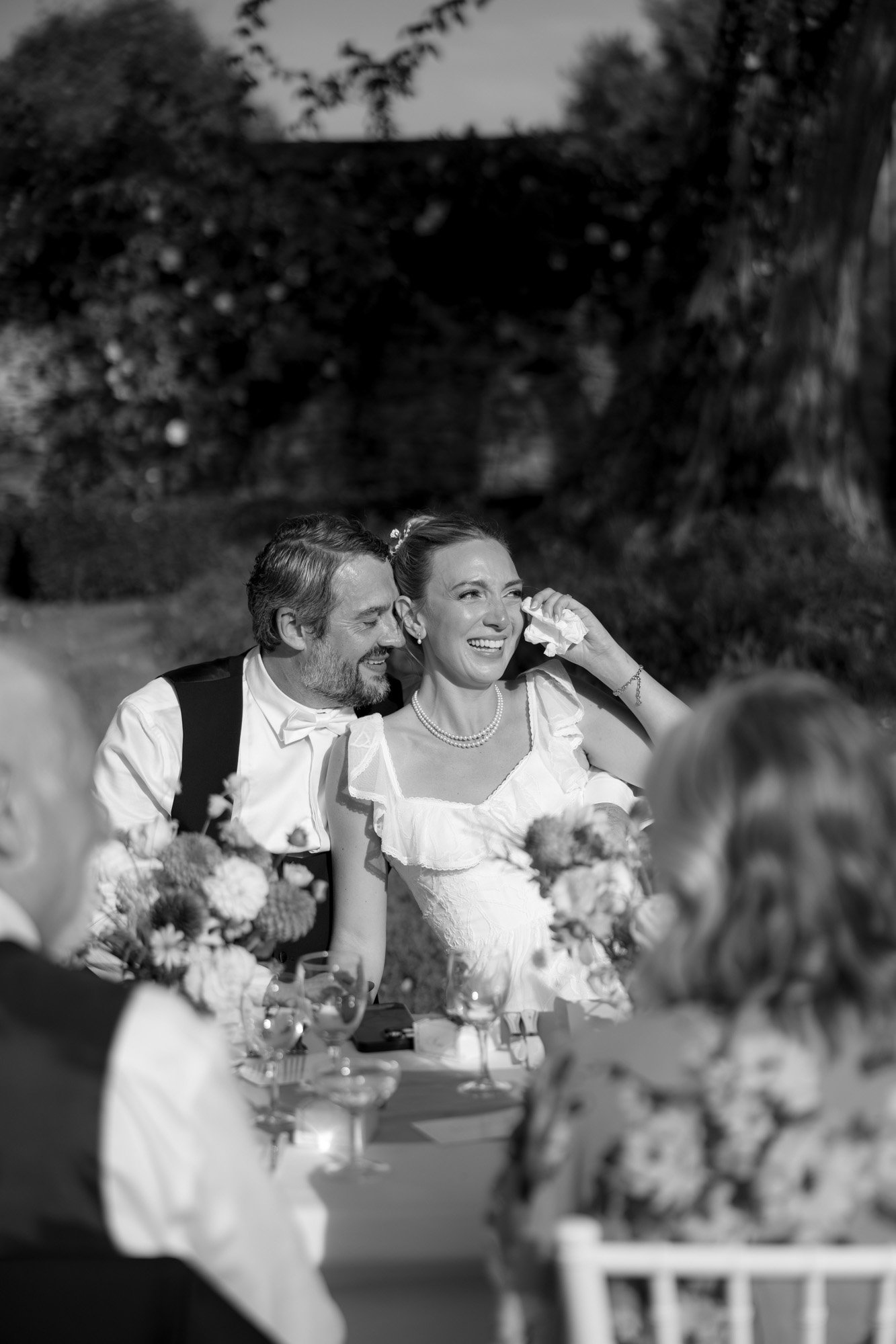 A bride wipes a tear while sitting next to a smiling man at an outdoor table; floral centerpieces and guests are visible in the foreground. Autumn France wedding.