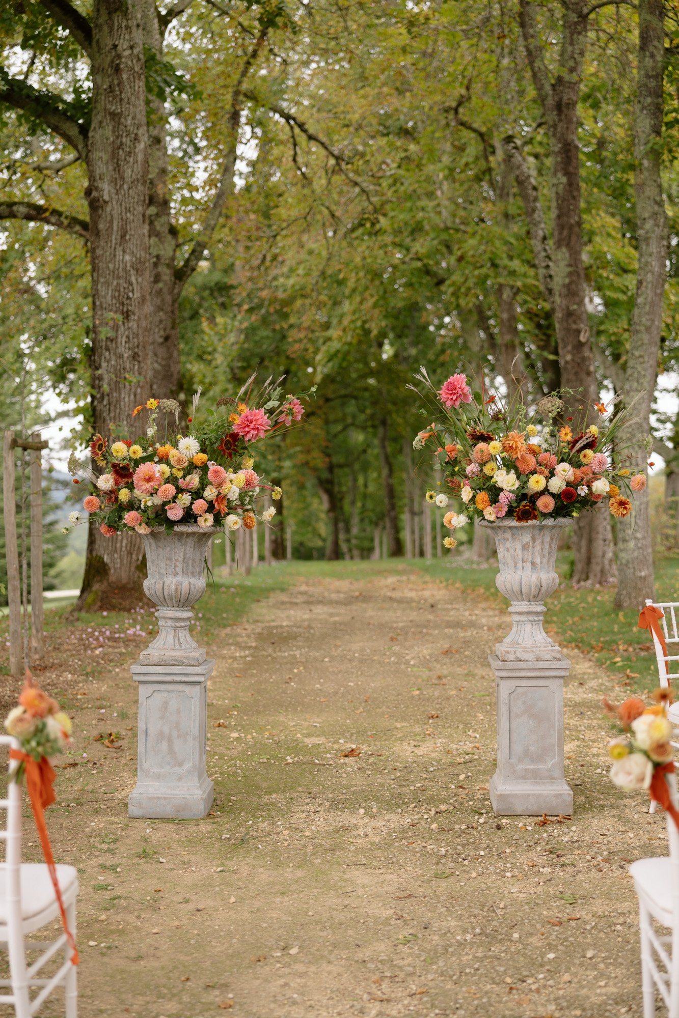 Two large stone urns filled with orange, pink, and yellow flowers stand on either side of a tree-lined outdoor path, with white chairs arranged along the sides. Autumn France wedding.