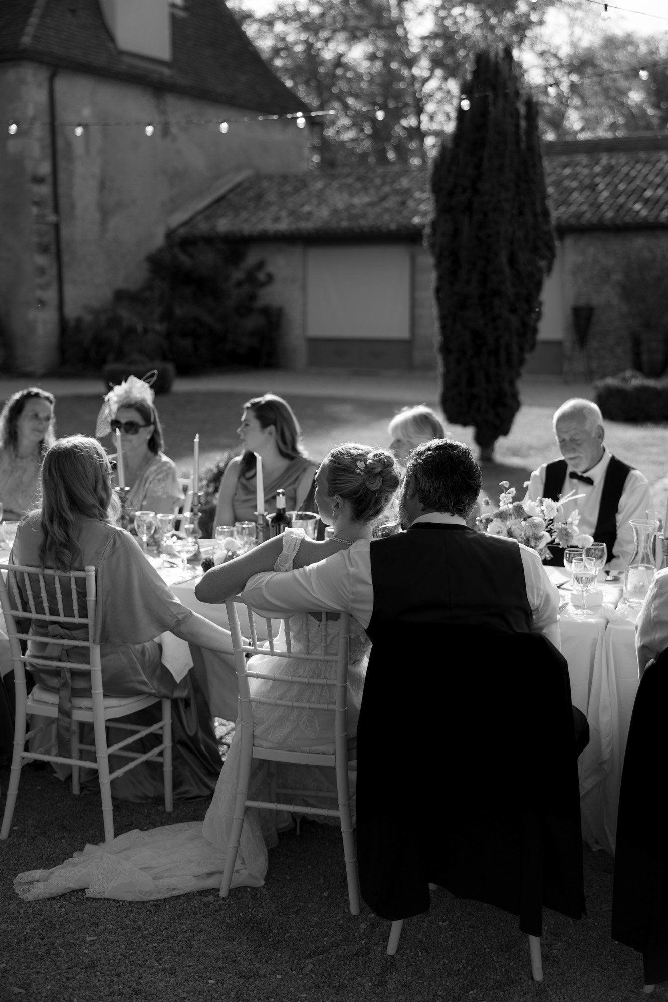 A group of people dressed formally sit around a long outdoor dining table set with candles and glassware during daylight.