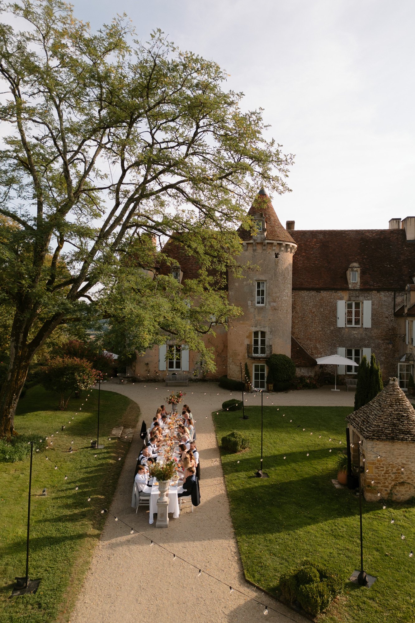 A long outdoor dining table is set for a group of people beneath a large tree in the courtyard of an old stone building with towers.