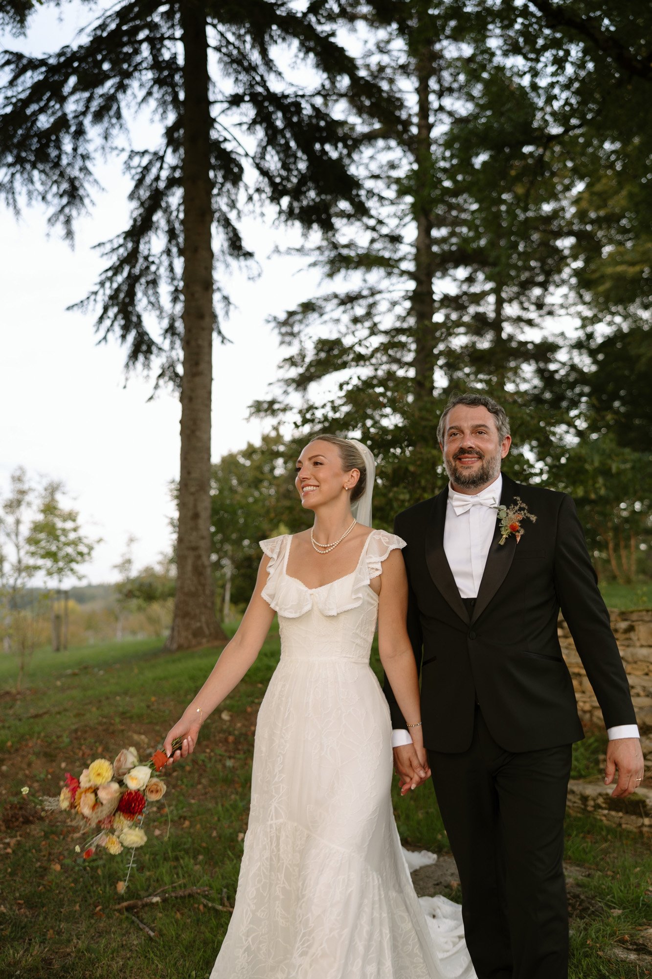 A bride in a white dress holding a bouquet and a groom in a black suit walk hand in hand outdoors, surrounded by trees. Autumn France wedding.