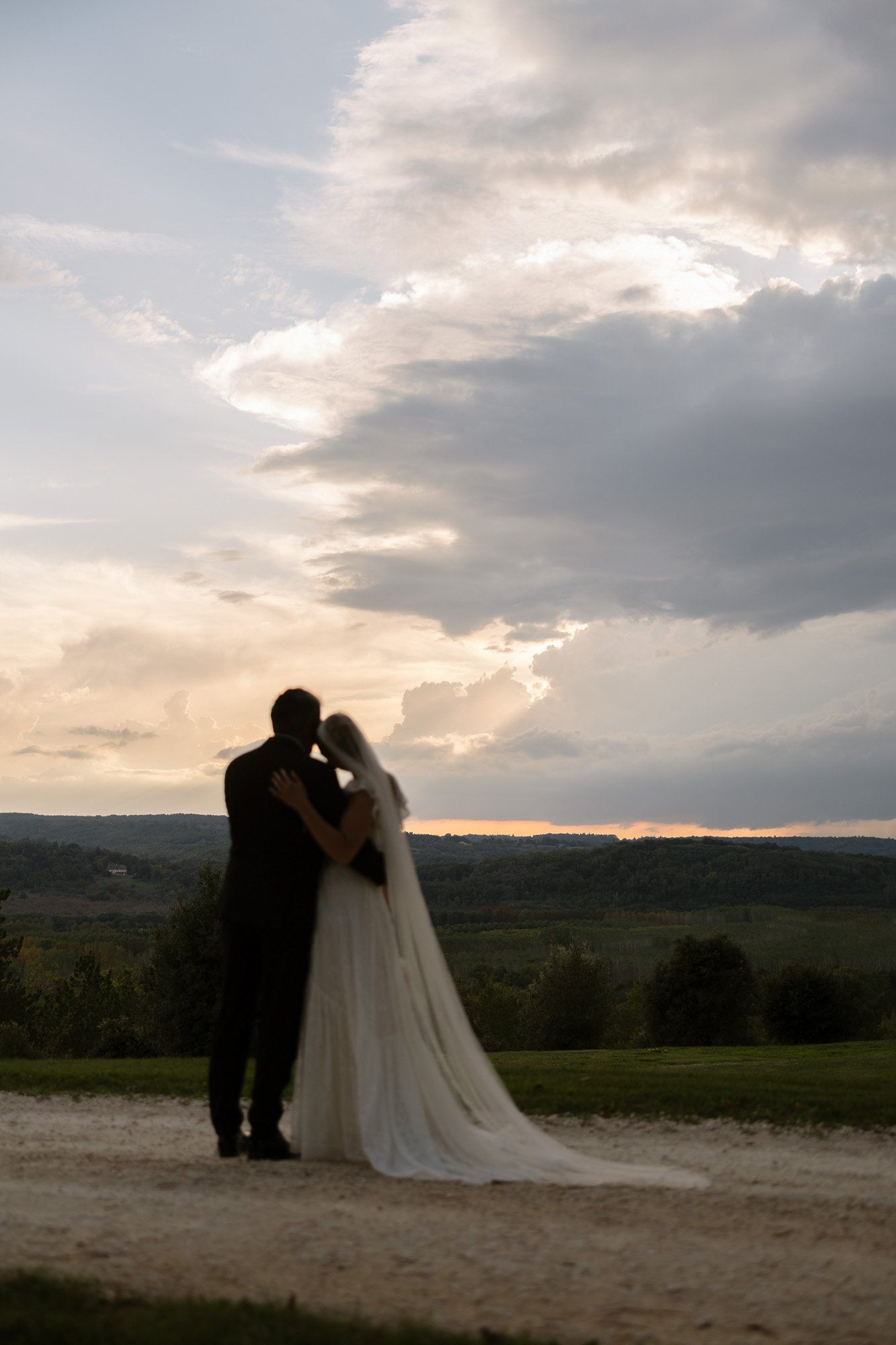 A bride and groom stand close together outdoors at sunset, facing each other, with a dramatic cloudy sky and landscape in the background. Autumn France wedding.